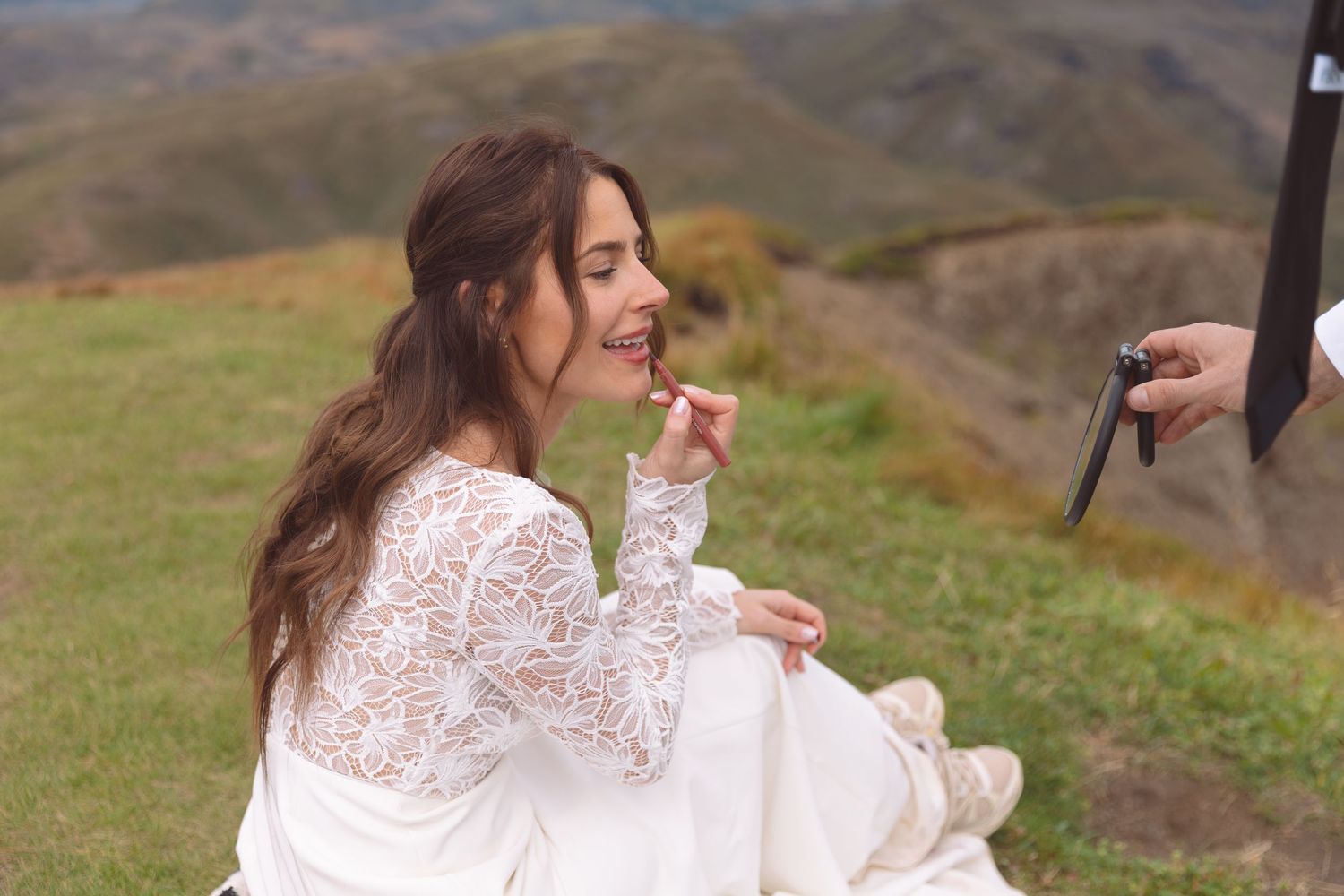 Smiling bride in white lace dress sits on hillside while groom holds mirror during wedding shoot.
