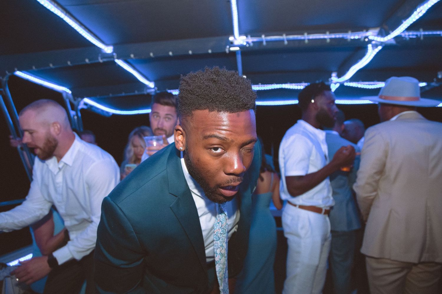 Young man in teal suit leaning forward at a lively wedding reception dance party.