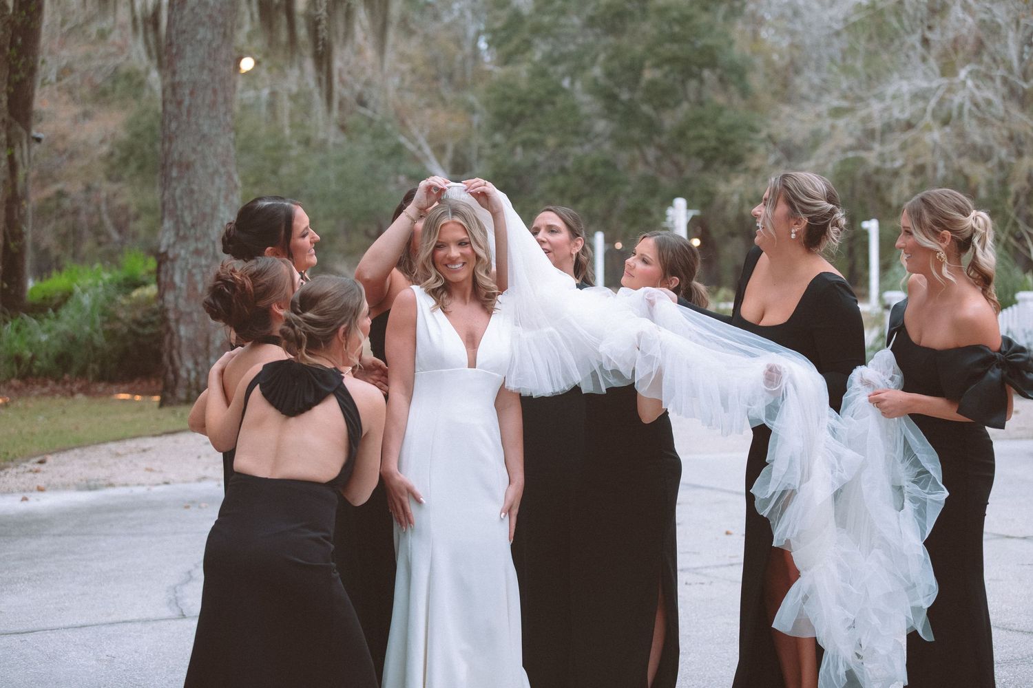 Bride in white gown celebrated by bridesmaids in black dresses lifting her veil outdoors.