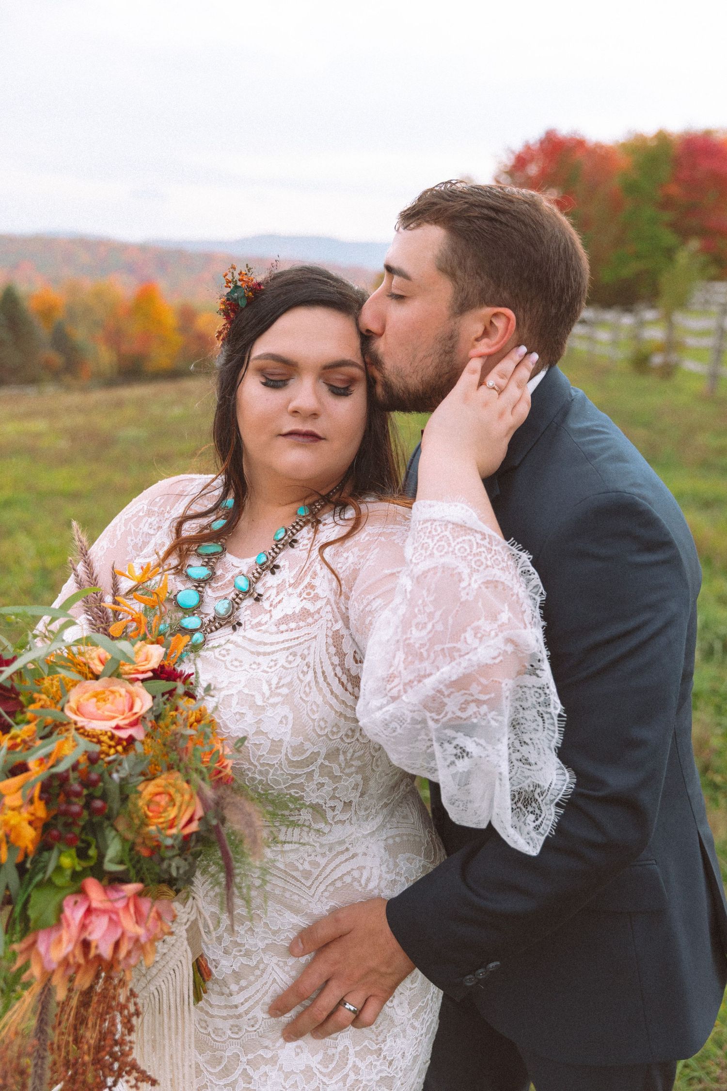 Groom kissing bride on cheek during fall outdoor wedding with colorful autumn foliage backdrop.