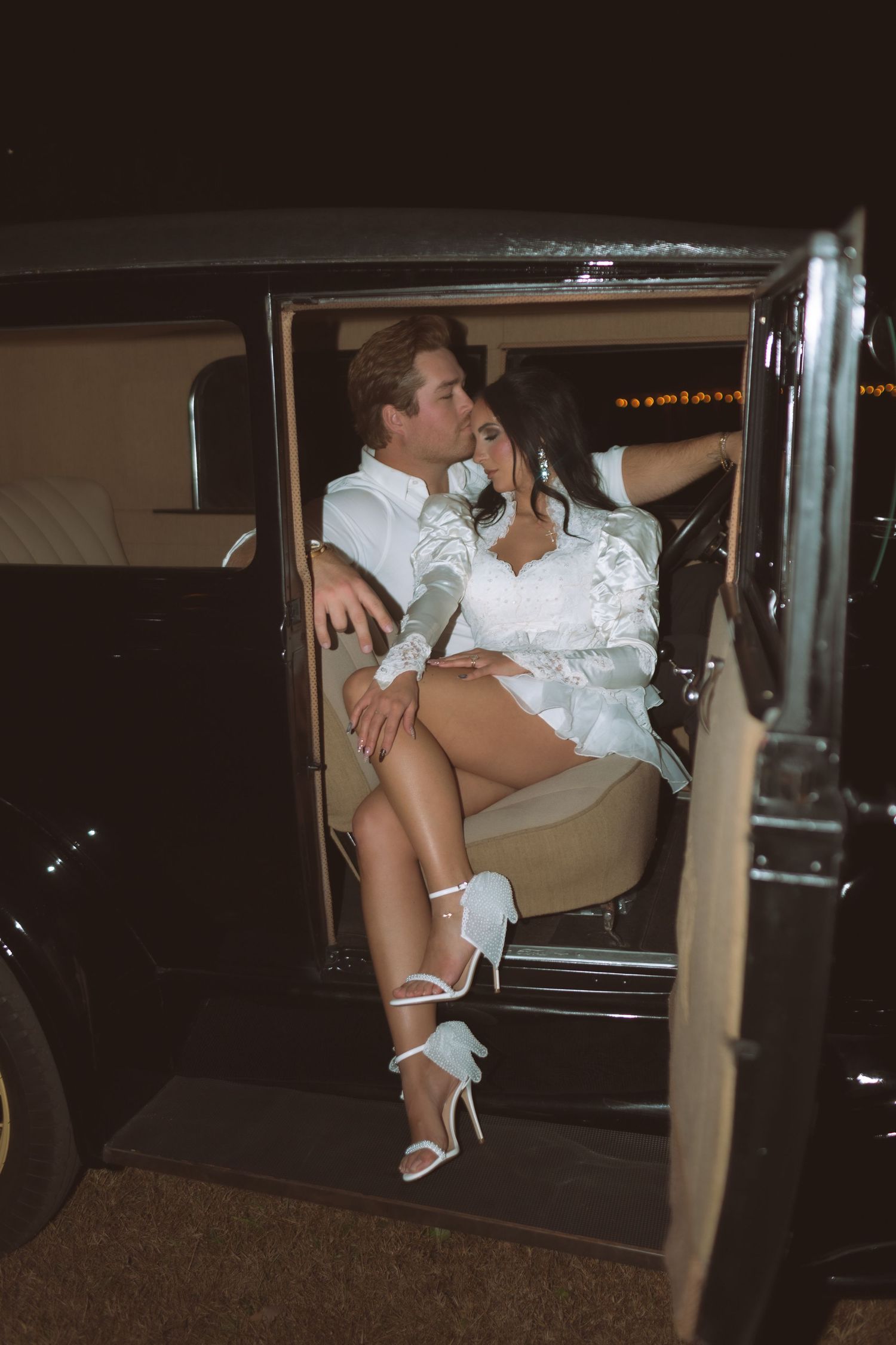 Bride and groom share a romantic moment inside a vintage classic car at night.