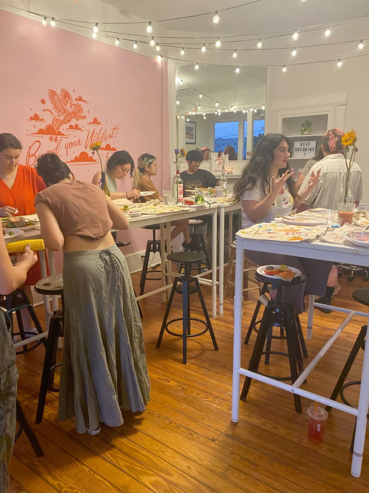 Art studio class with people painting at high tables under string lights with a pink accent wall.