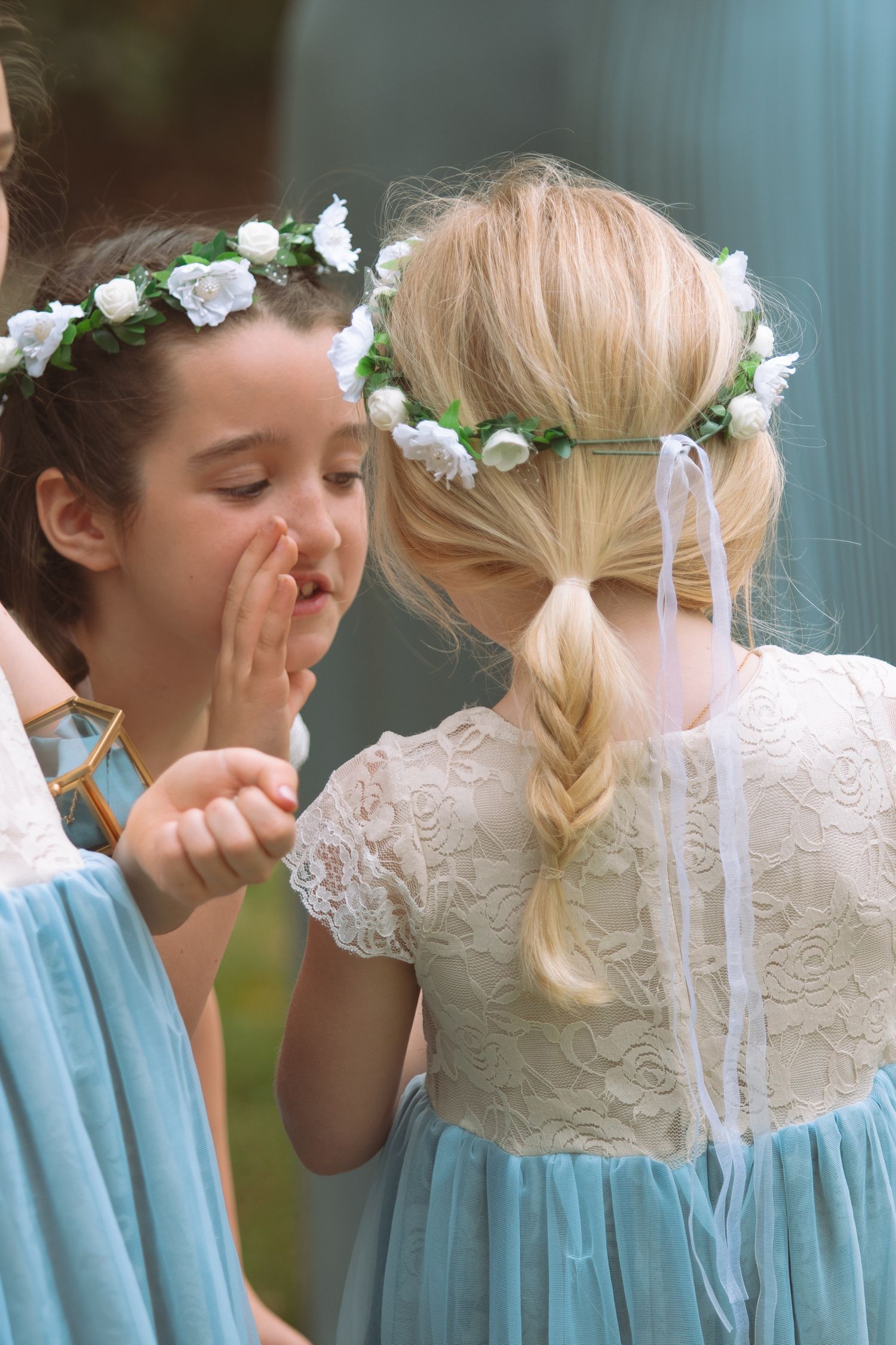 Two young flower girls in floral crowns and blue dresses share a secret at wedding.