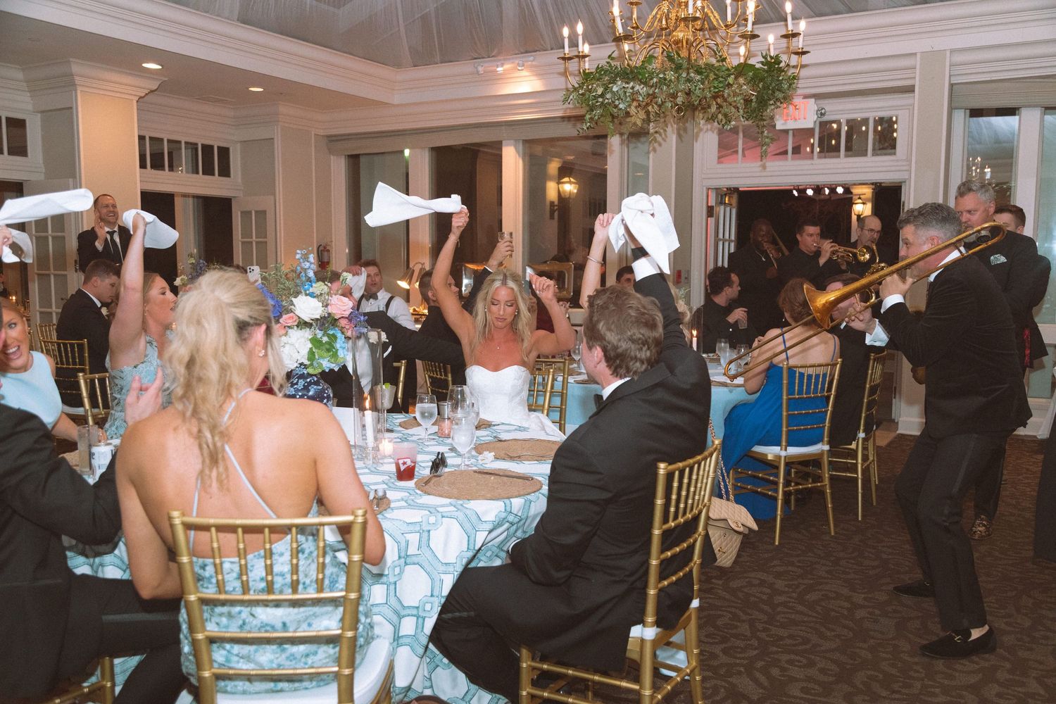Wedding reception guests wave white napkins while a brass band performs in an elegant ballroom with chandeliers.