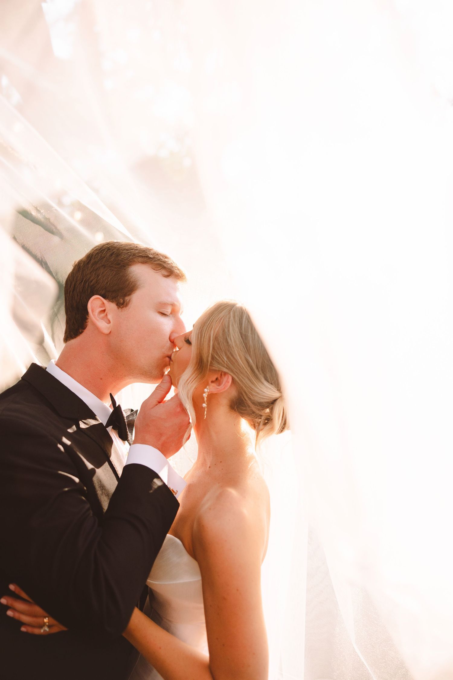 A groom in a black tuxedo tenderly kissing his bride wrapped in a flowing veil in golden light.