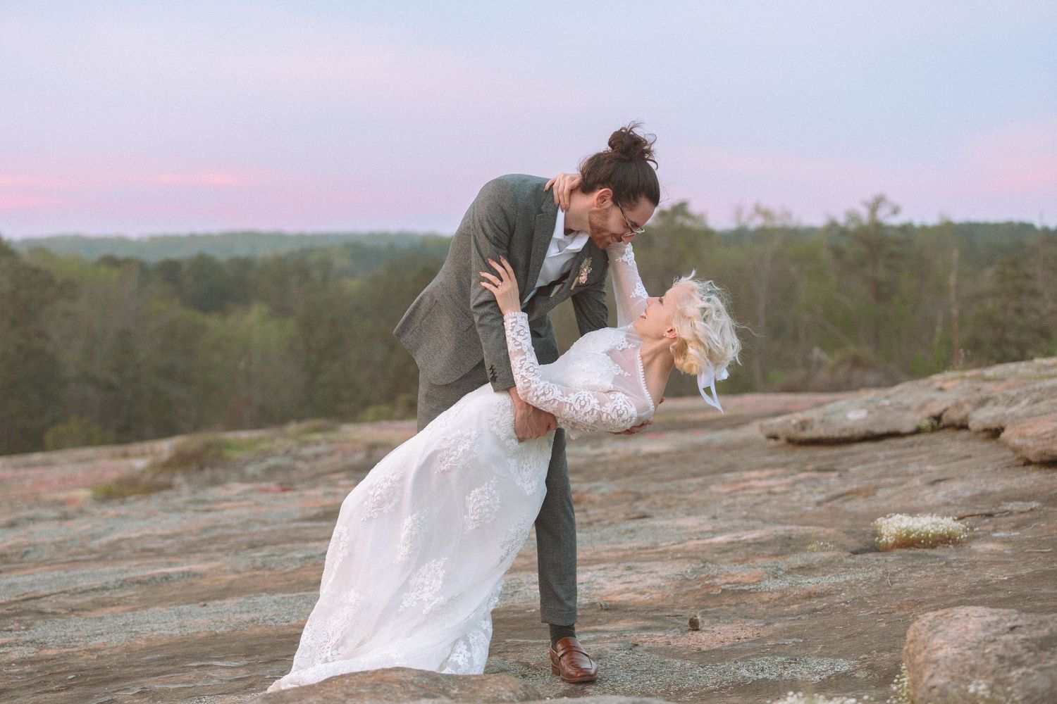 Romantic wedding couple sharing a dip kiss on rocky terrain at sunset with pink sky backdrop.