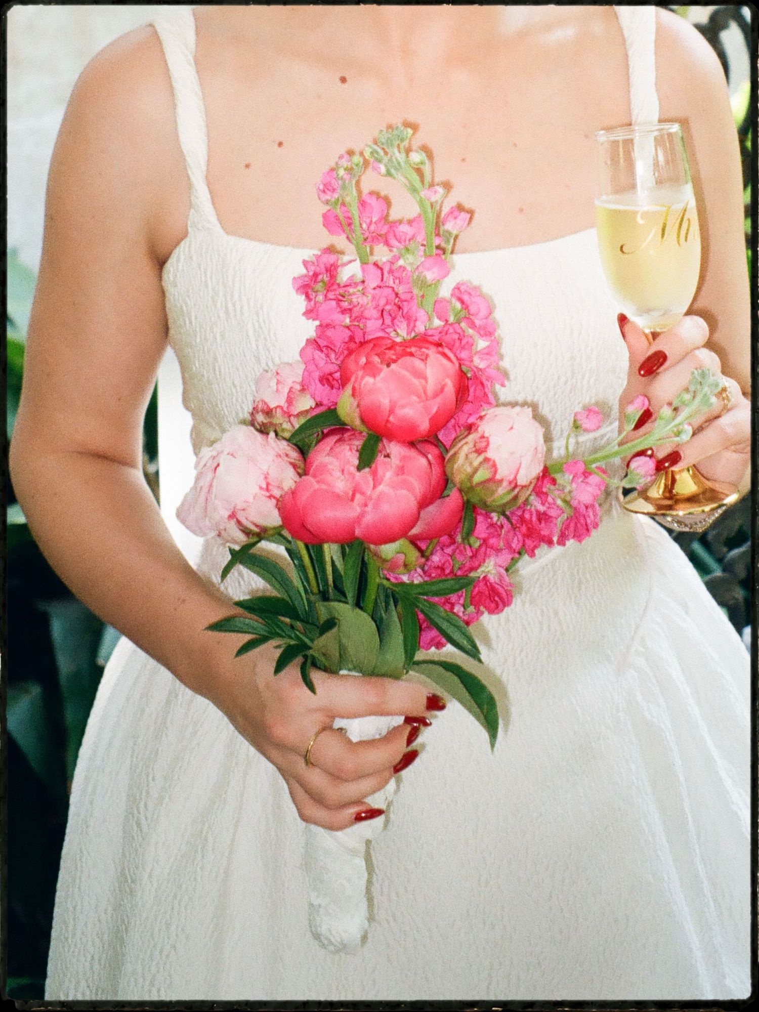 Bride in white dress holding a vibrant pink peony bouquet and a glass of champagne outdoors.