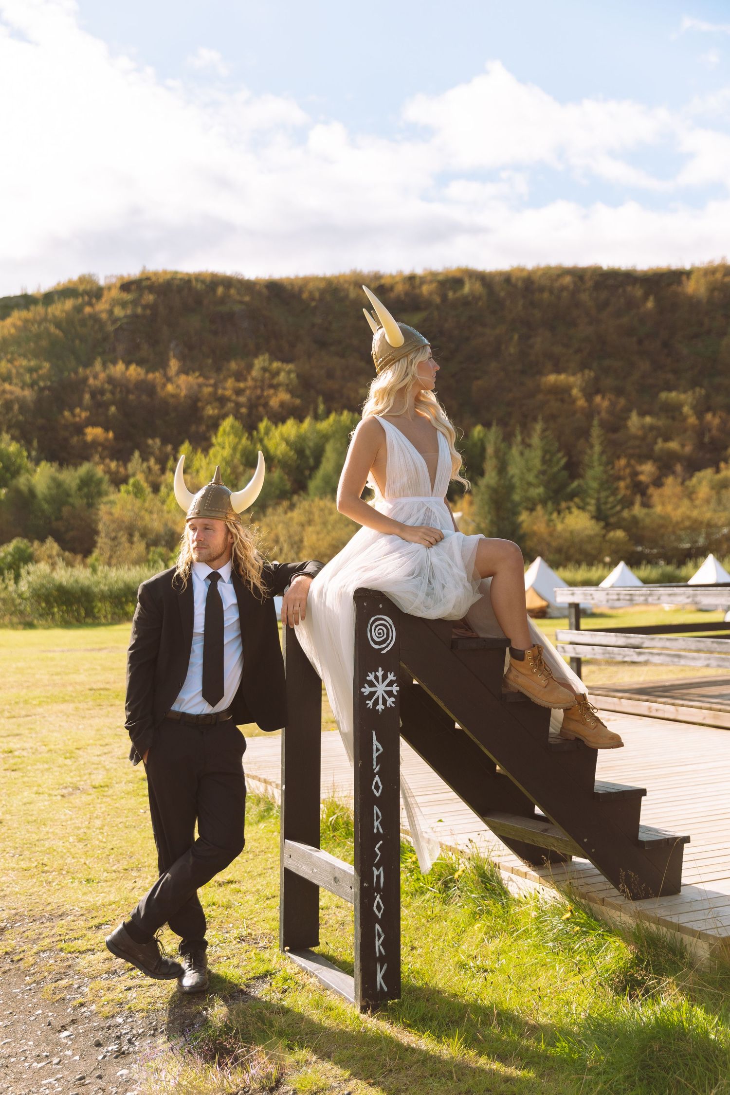 Viking-themed wedding couple pose on rustic wooden stairs outdoors with scenic green hills backdrop.