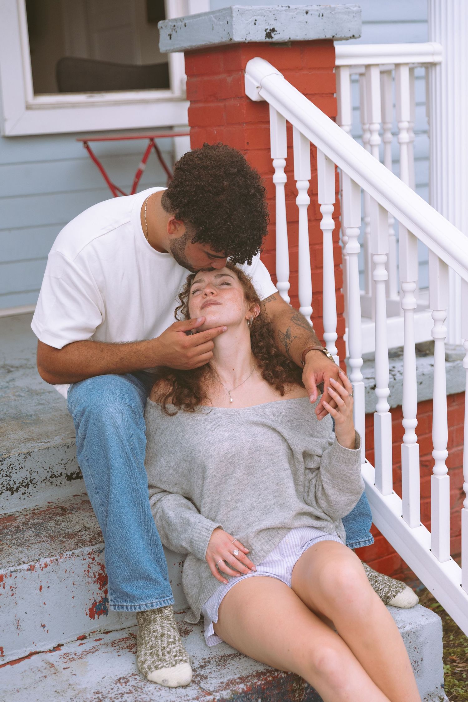 Affectionate couple sitting on porch steps, man gently kisses woman in gray sweater on the cheek.