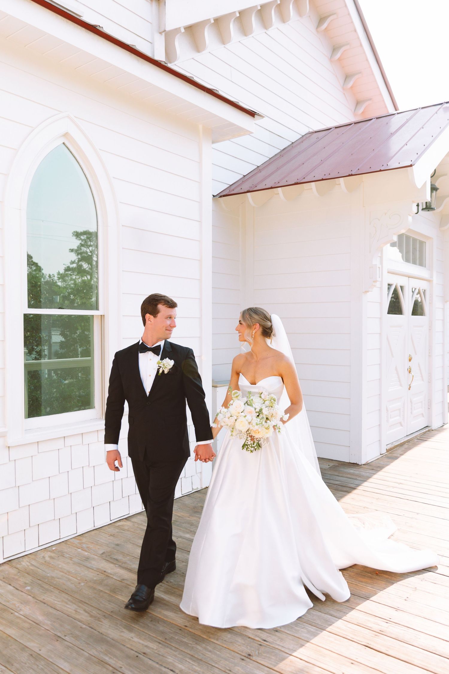 Bride and groom walk hand in hand outside white chapel venue, bride holding white bouquet in golden light.