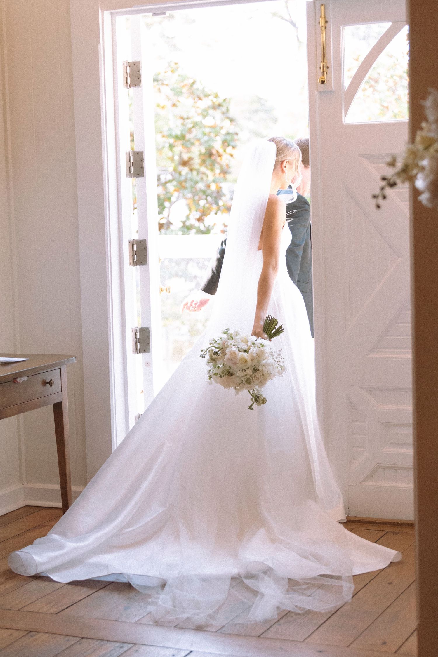 Bride in flowing white gown with veil holding bouquet stands at open white door with groom.