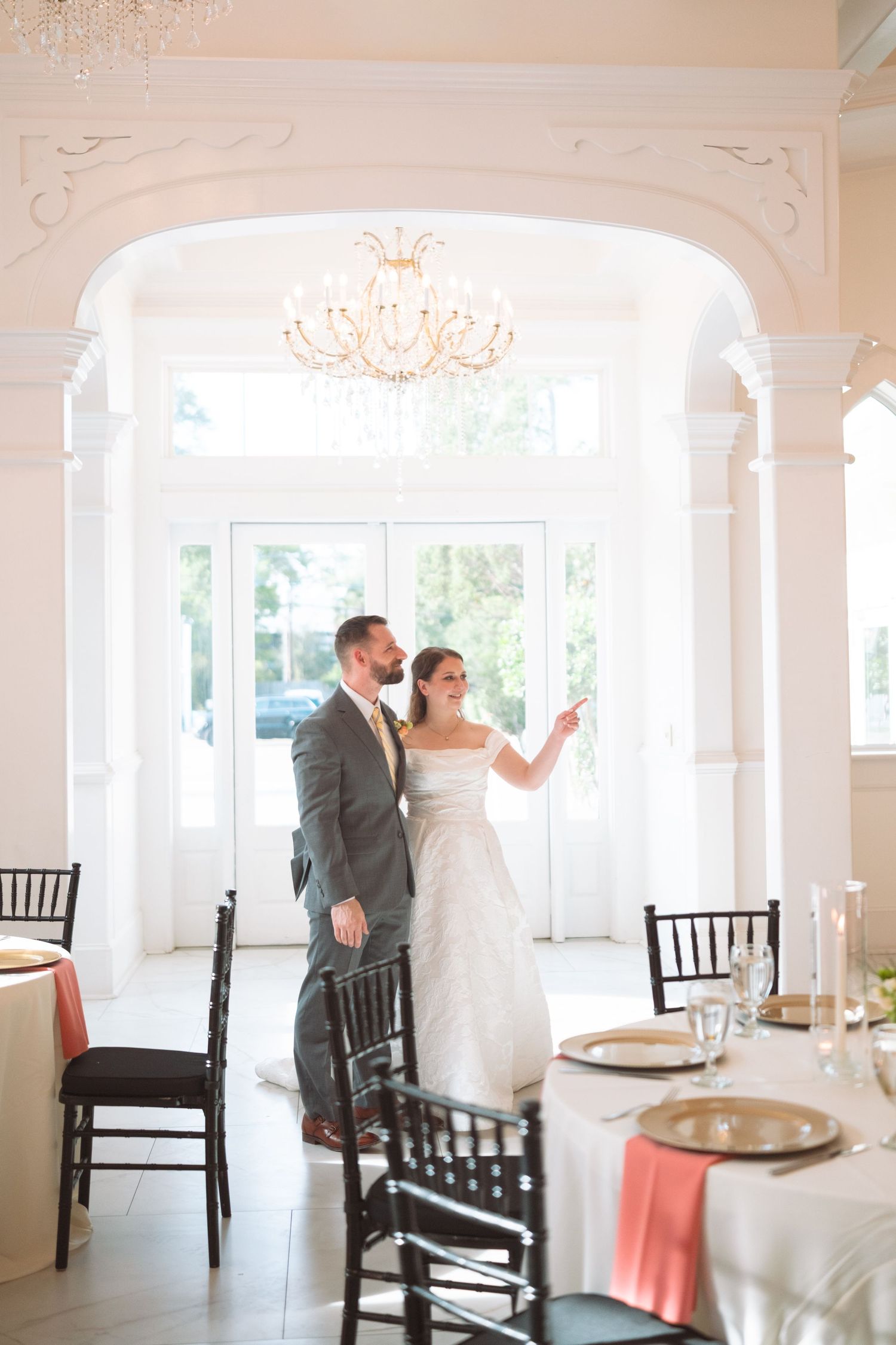 Bride and groom share a romantic moment in an elegant white ballroom with chandelier and coral-accented reception tables.