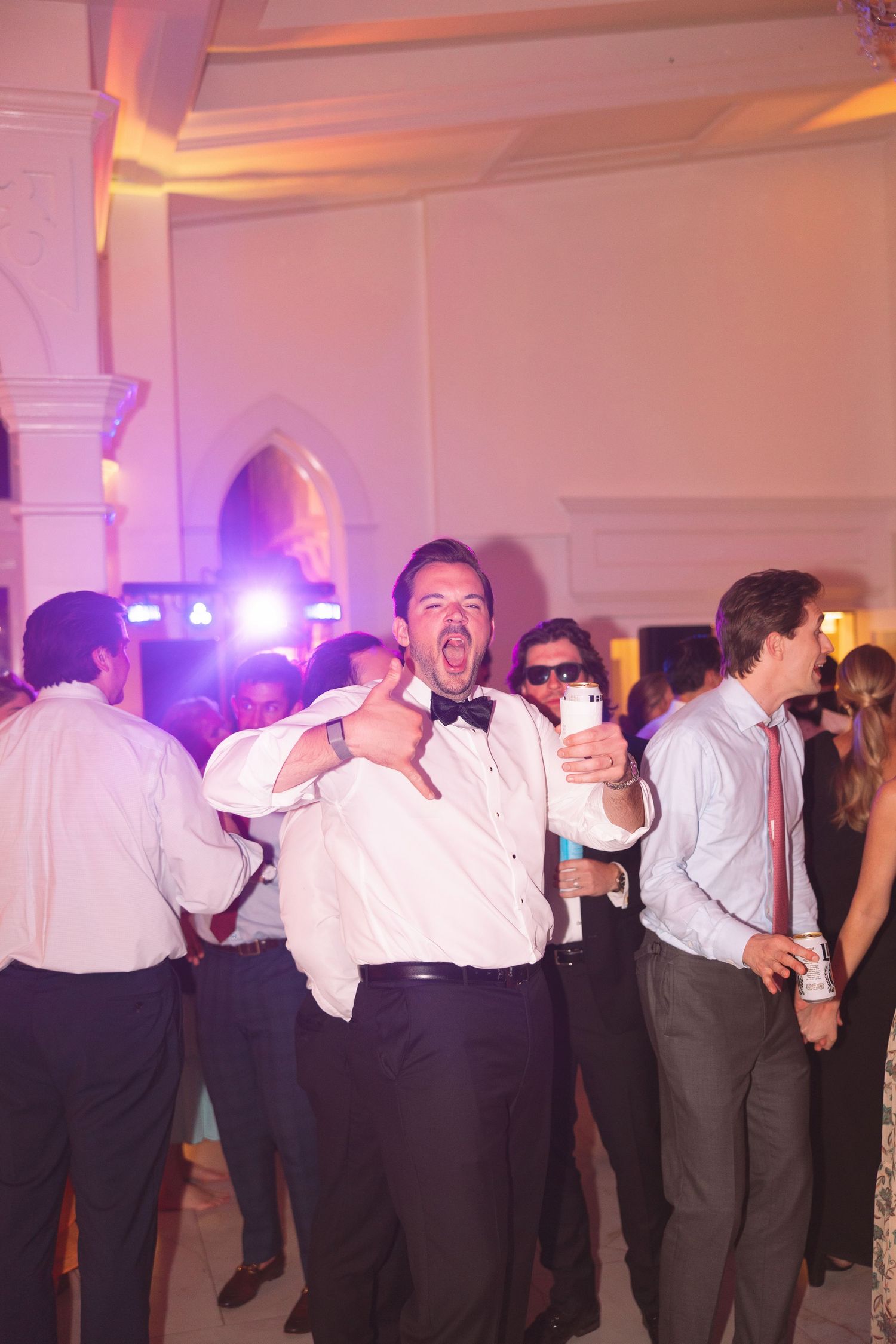 Excited man in bow tie giving thumbs up at elegant wedding reception party with colorful DJ lights.