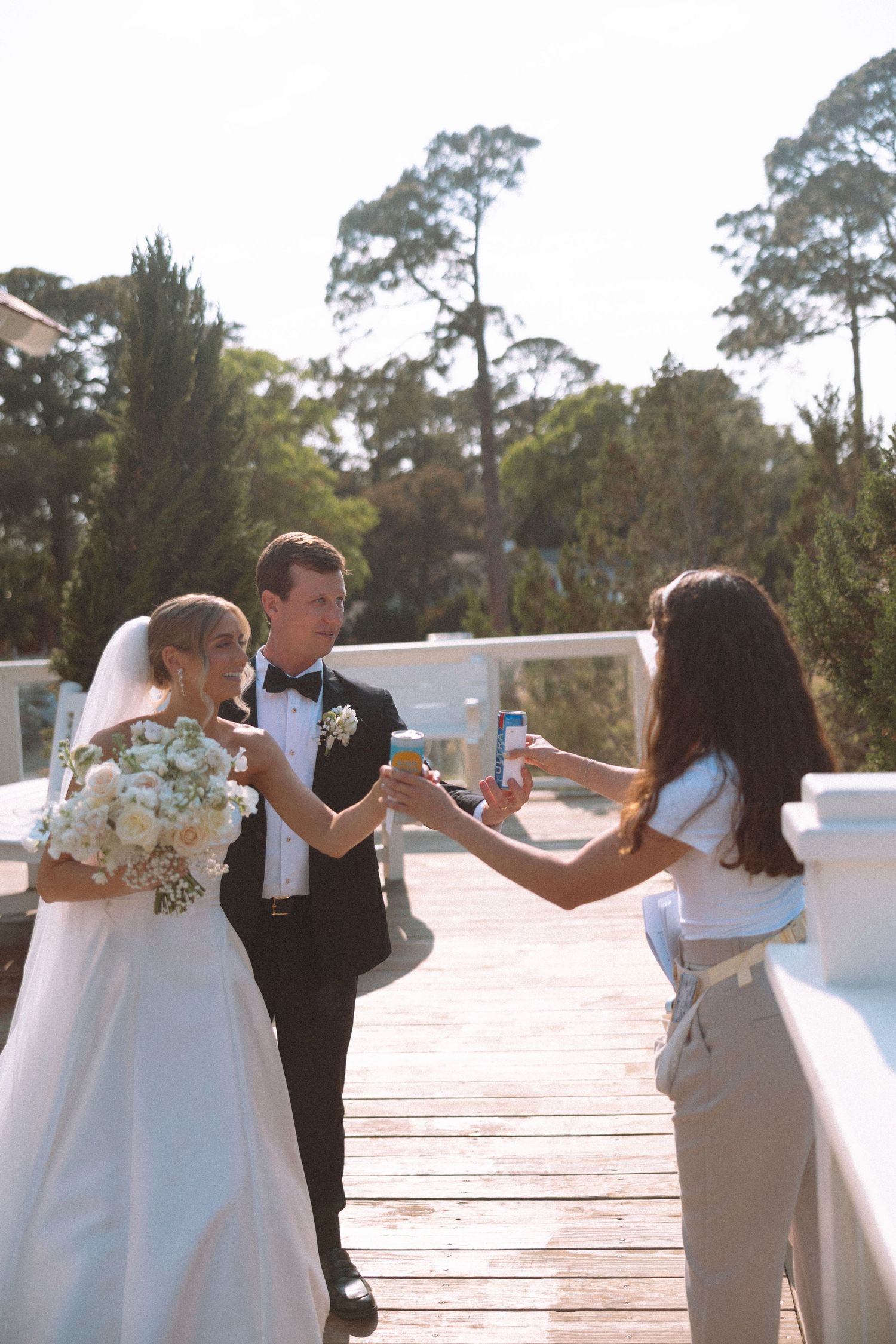Bride and groom toasting champagne with a guest on a sunny outdoor deck surrounded by trees.