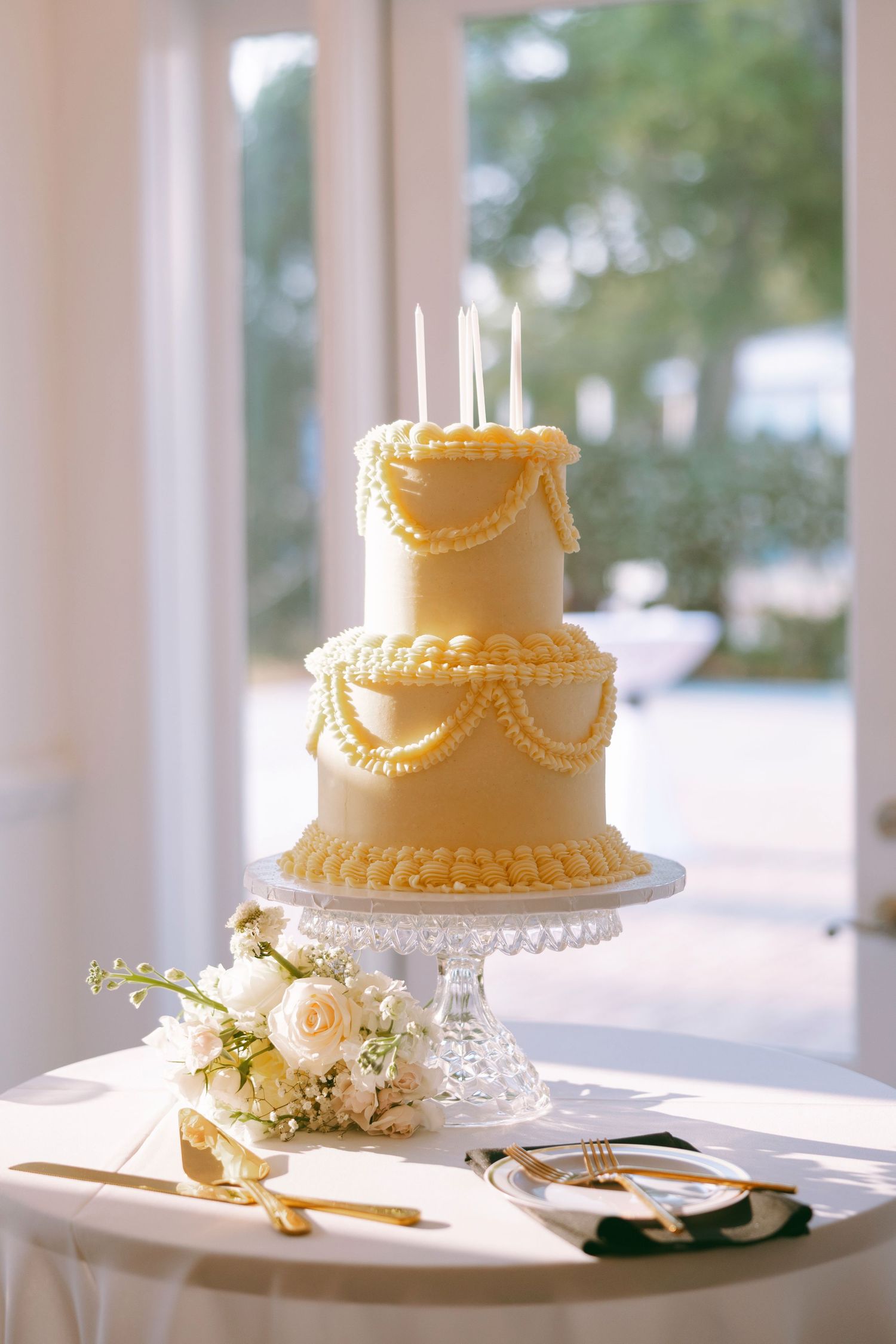 Elegant two-tier yellow wedding cake with pearl swag details and candles on crystal stand beside white roses.