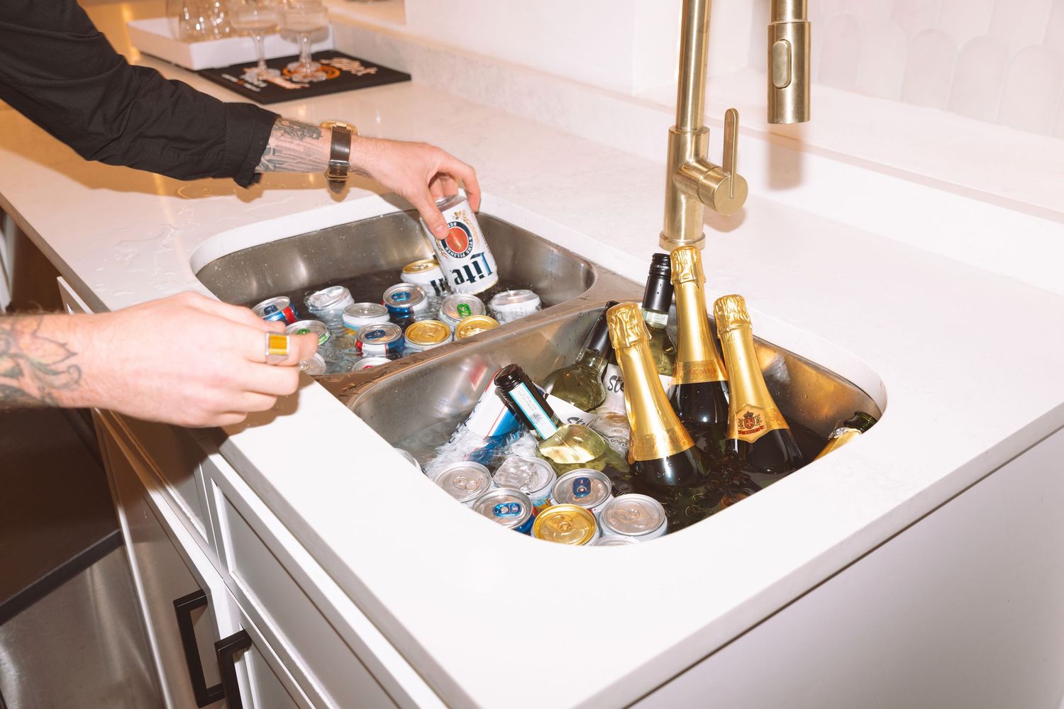 Person grabbing canned drinks from an ice-filled kitchen sink next to champagne bottles at a party.