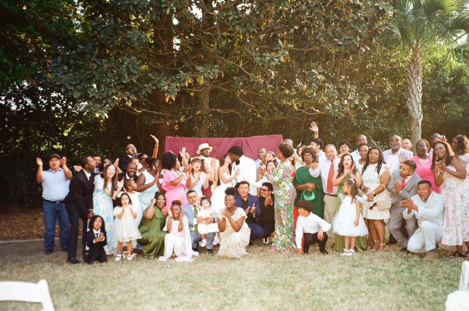 Large diverse group of wedding guests posing together outdoors under trees at a backyard celebration.