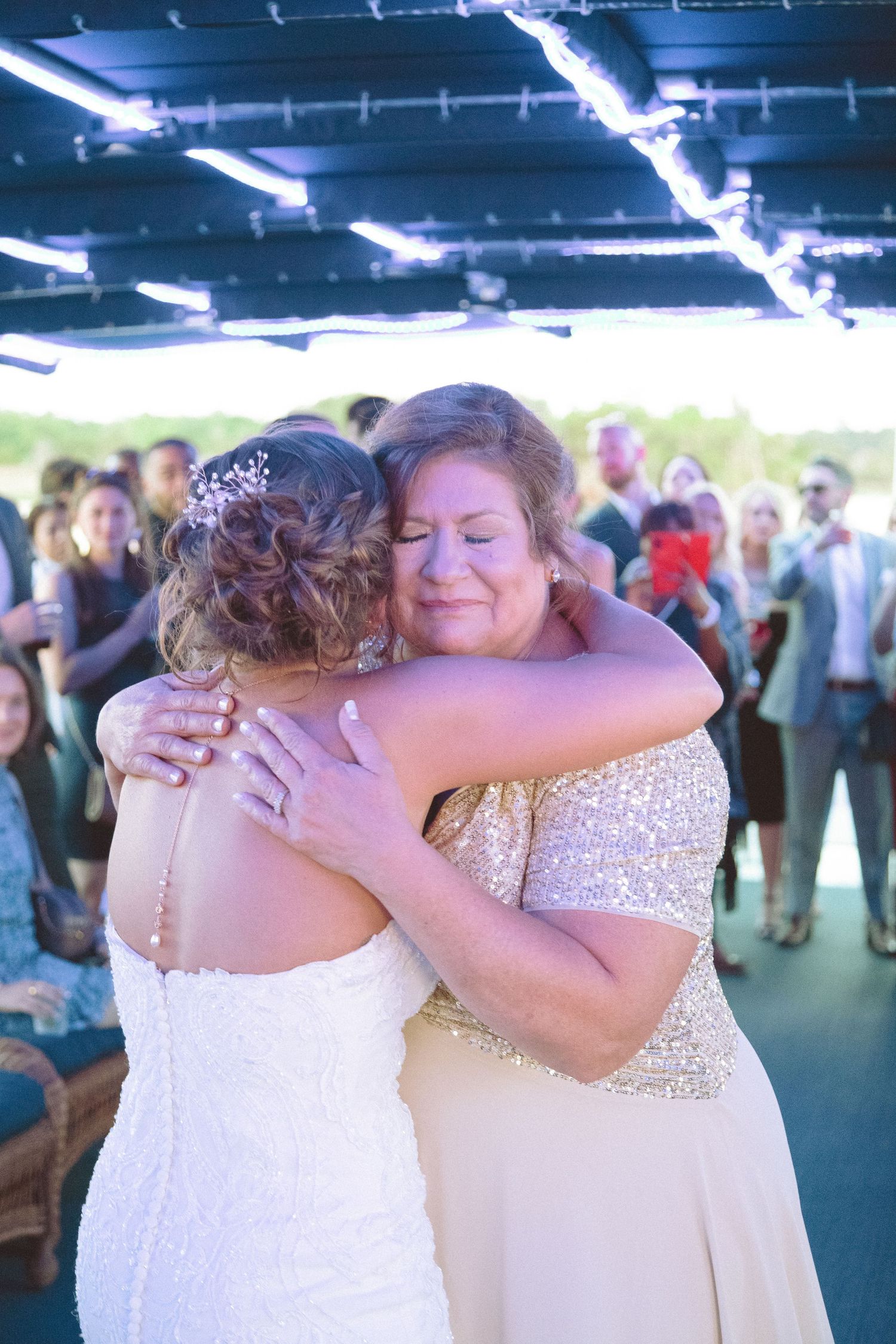 Emotional mother hugs bride in white lace gown at wedding reception on a boat.