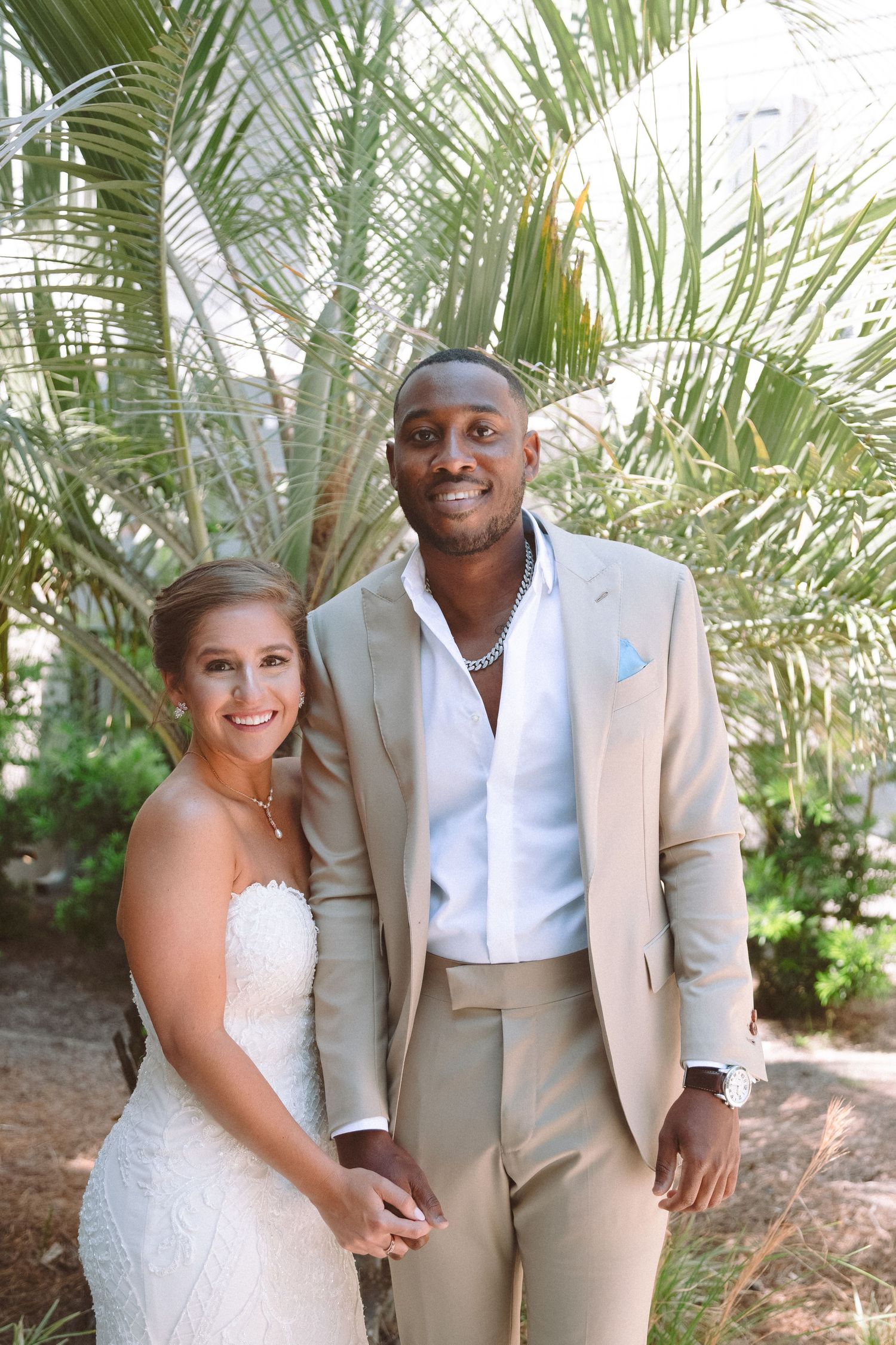 Interracial couple posing together outdoors in wedding attire with palm trees in background.