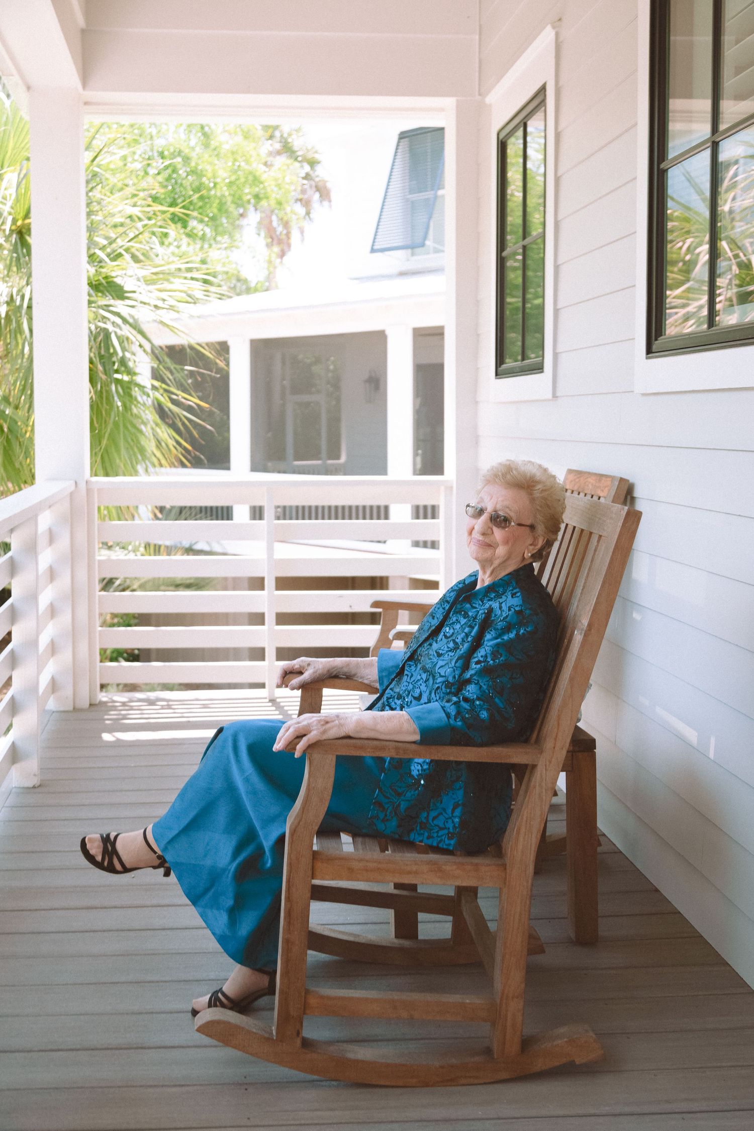 Elegant elderly woman in teal outfit relaxing in wooden rocking chair on a sunny white porch.