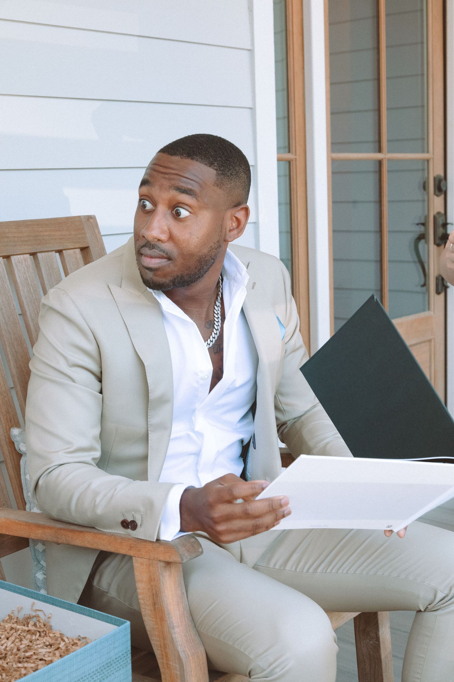 Well-dressed man in tan suit holding papers with thoughtful expression while sitting in wooden chair outside.