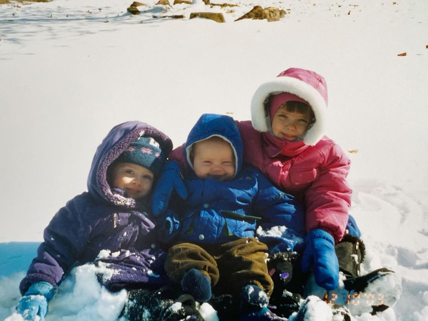 Three young children bundled in colorful winter snowsuits sitting together in the snow outdoors.
