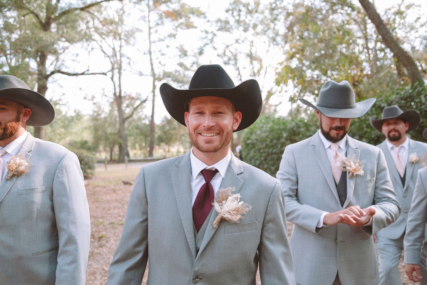 Groom and groomsmen in grey suits and black cowboy hats at rustic outdoor wedding.