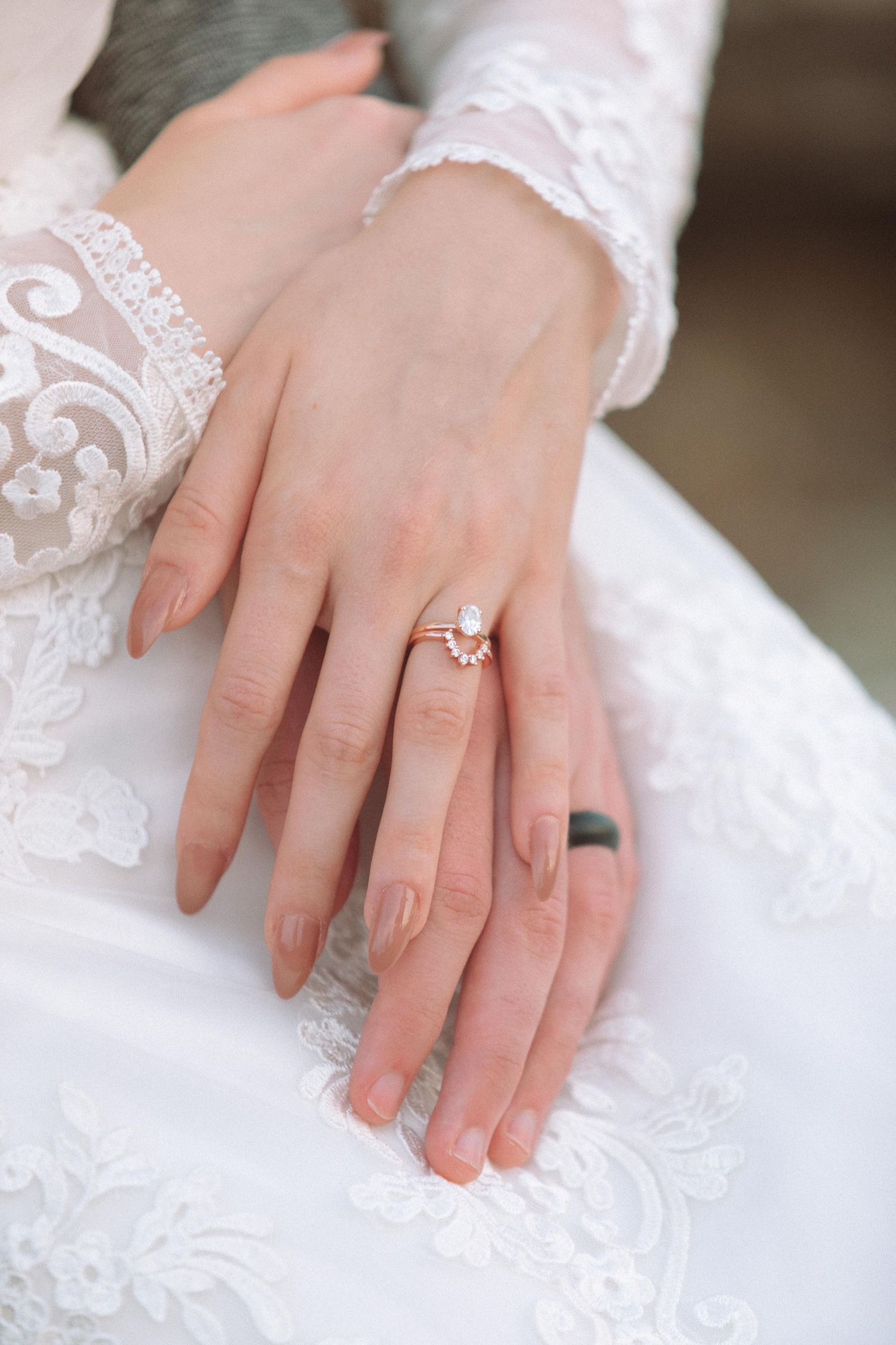 Close-up of bride's hands showing elegant pearl and gold engagement ring on white lace dress.