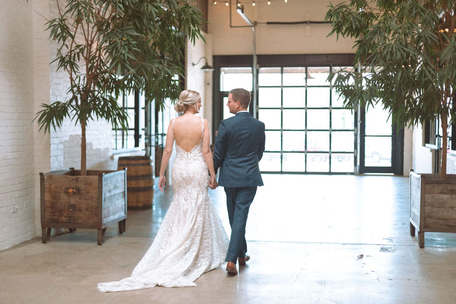 Bride and groom walk hand in hand through modern industrial wedding venue with bamboo trees.