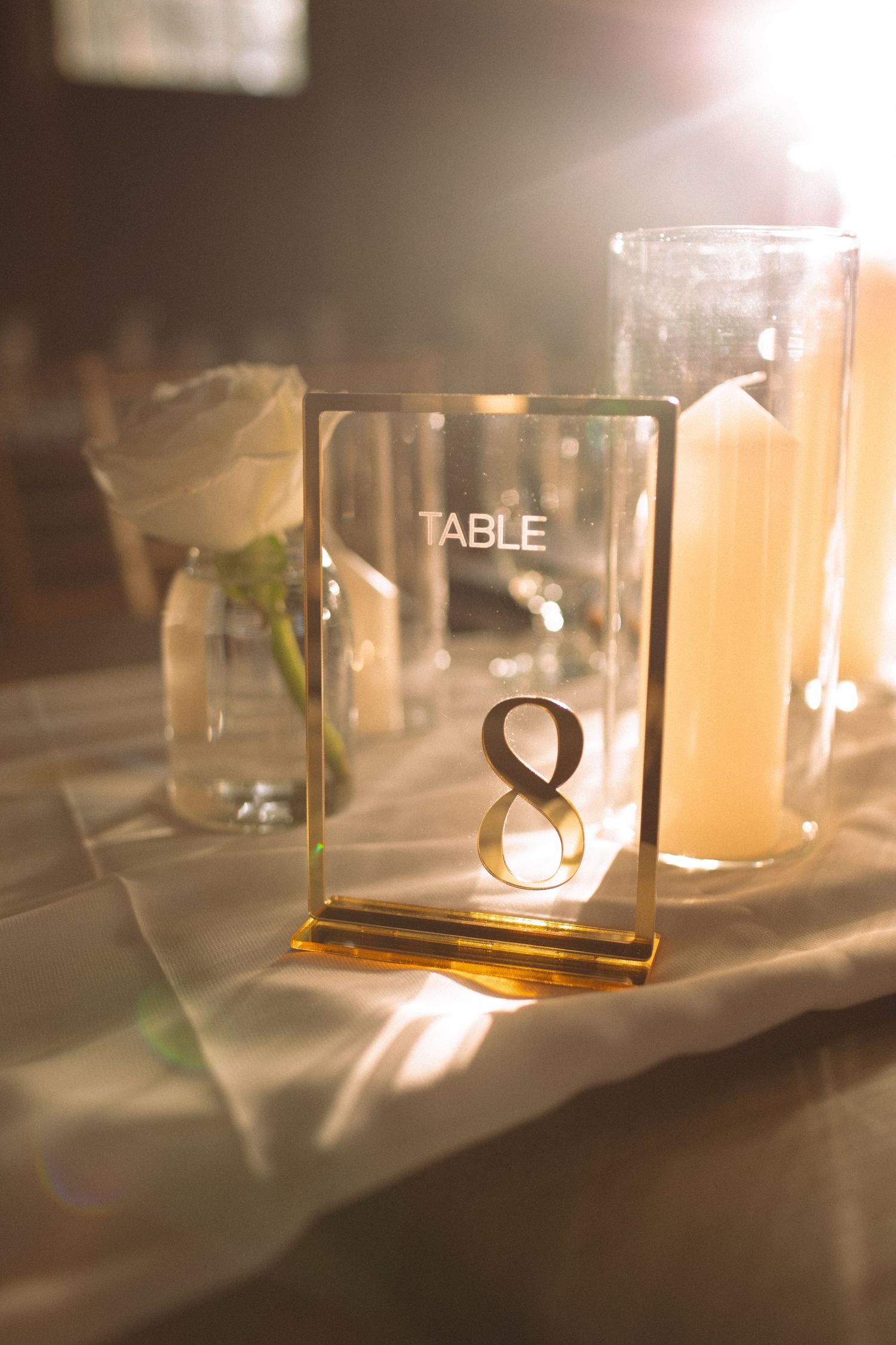 Gold acrylic table number 8 sign with candle and white rose centerpiece at elegant wedding reception.