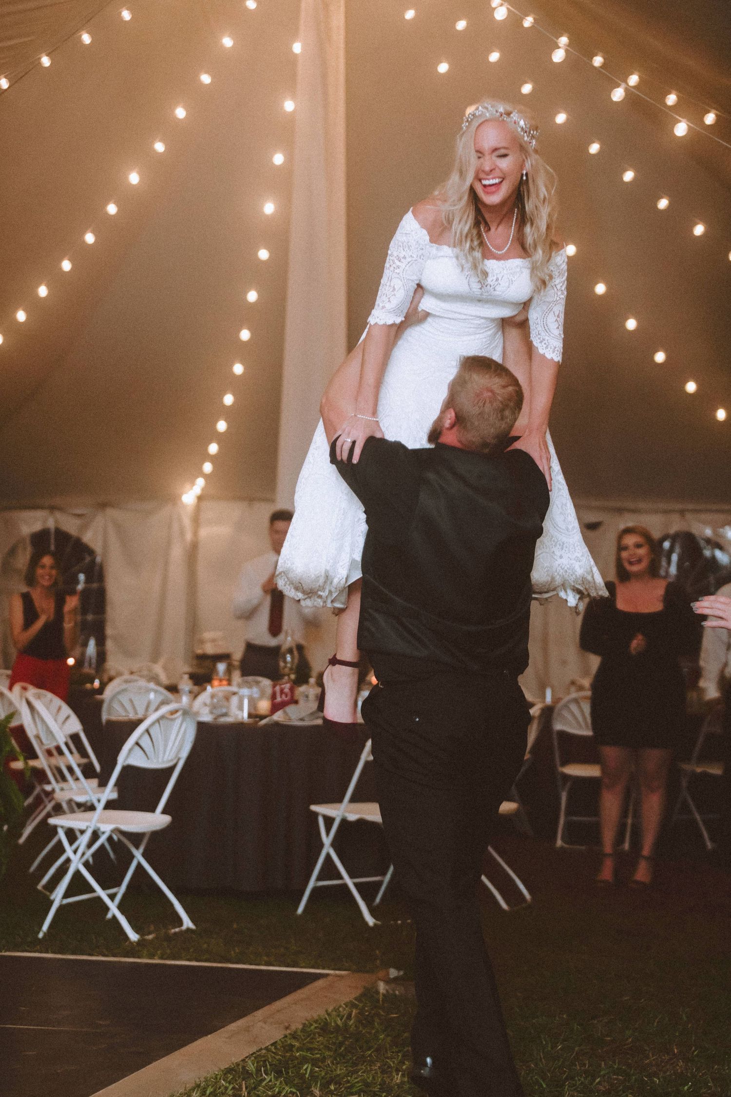Groom joyfully lifting laughing bride during wedding reception dance under warm string lights in tent.