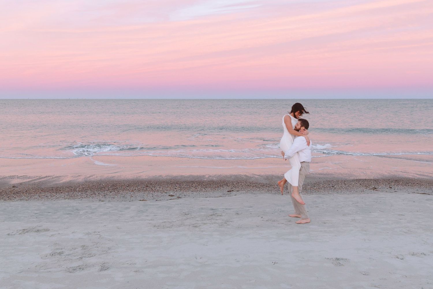 Couple embracing on a sandy beach at sunset with a pink and purple sky over calm ocean waters.