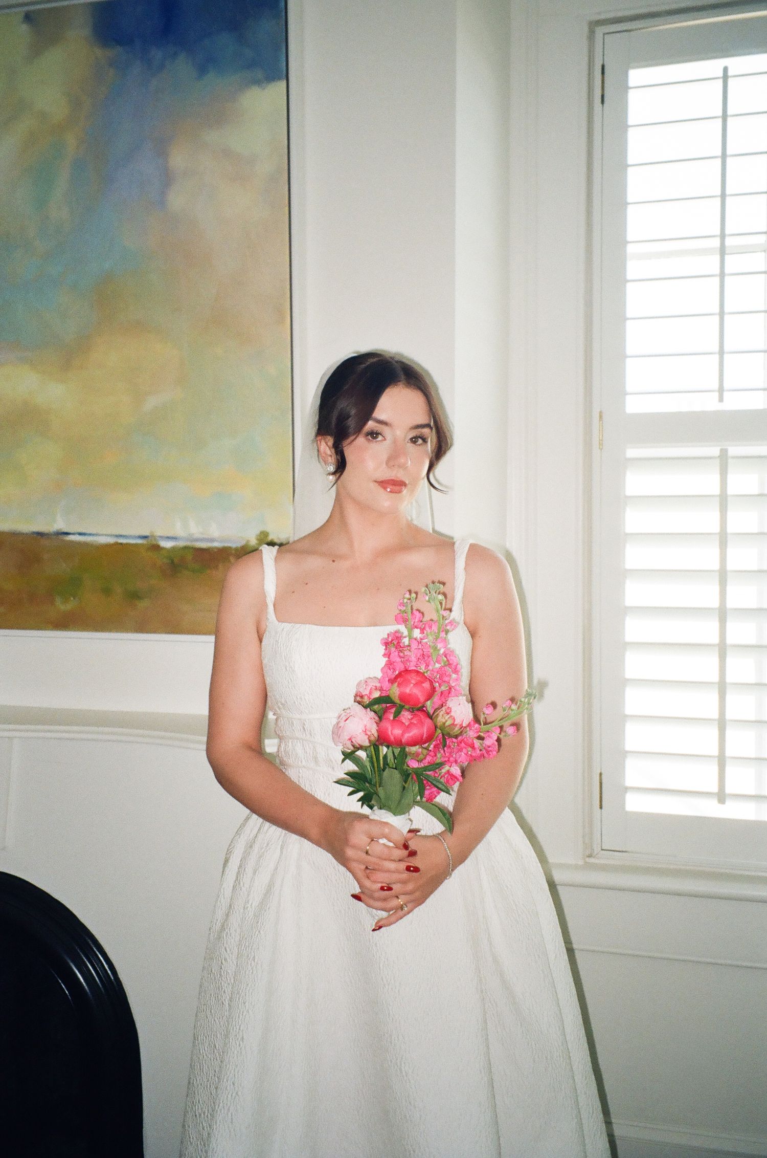 Bride in white dress holding pink peony bouquet stands by window with plantation shutters and abstract painting.