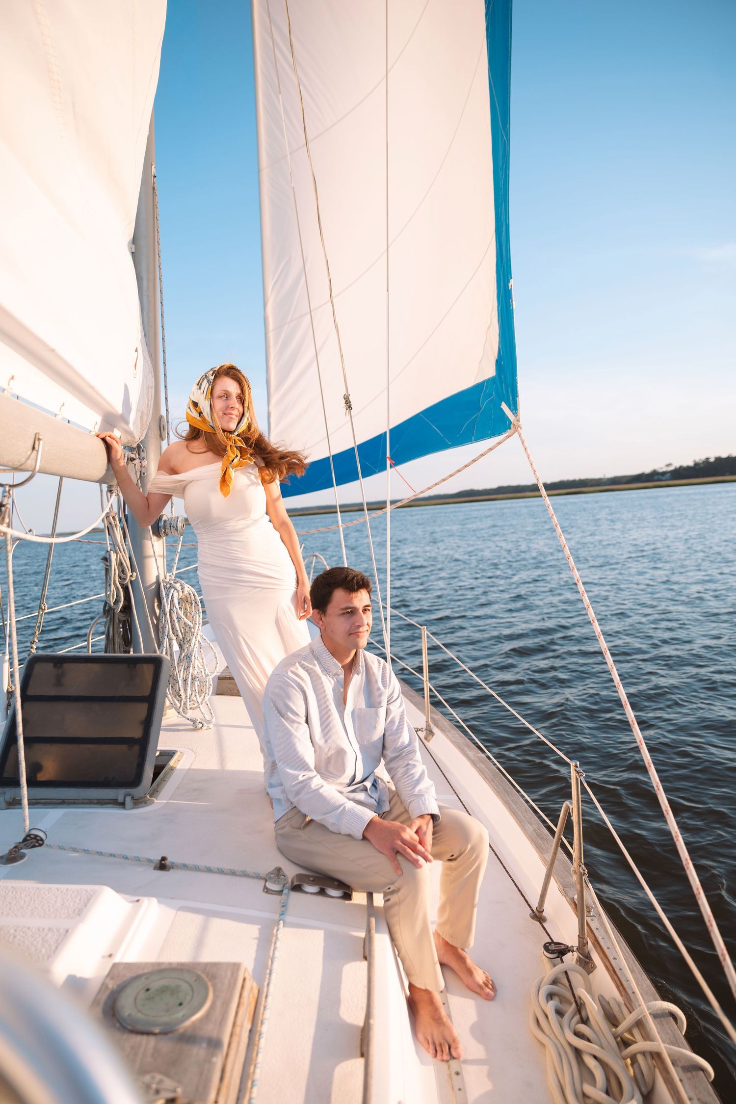 A woman in a white dress stands on a sailboat deck while a man sits beside her under a bright blue sky.