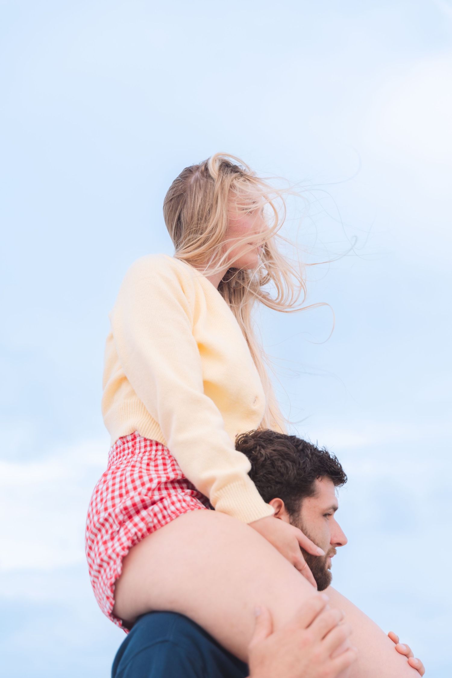 Woman sitting on mans shoulders laughing at the beach with wind in her hair.
