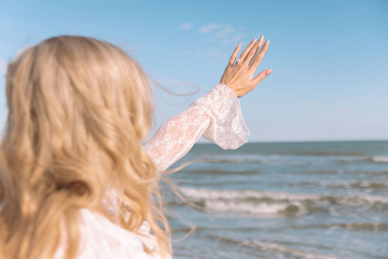 Blonde woman in white lace dress raising hand toward ocean waves on sunny beach day.
