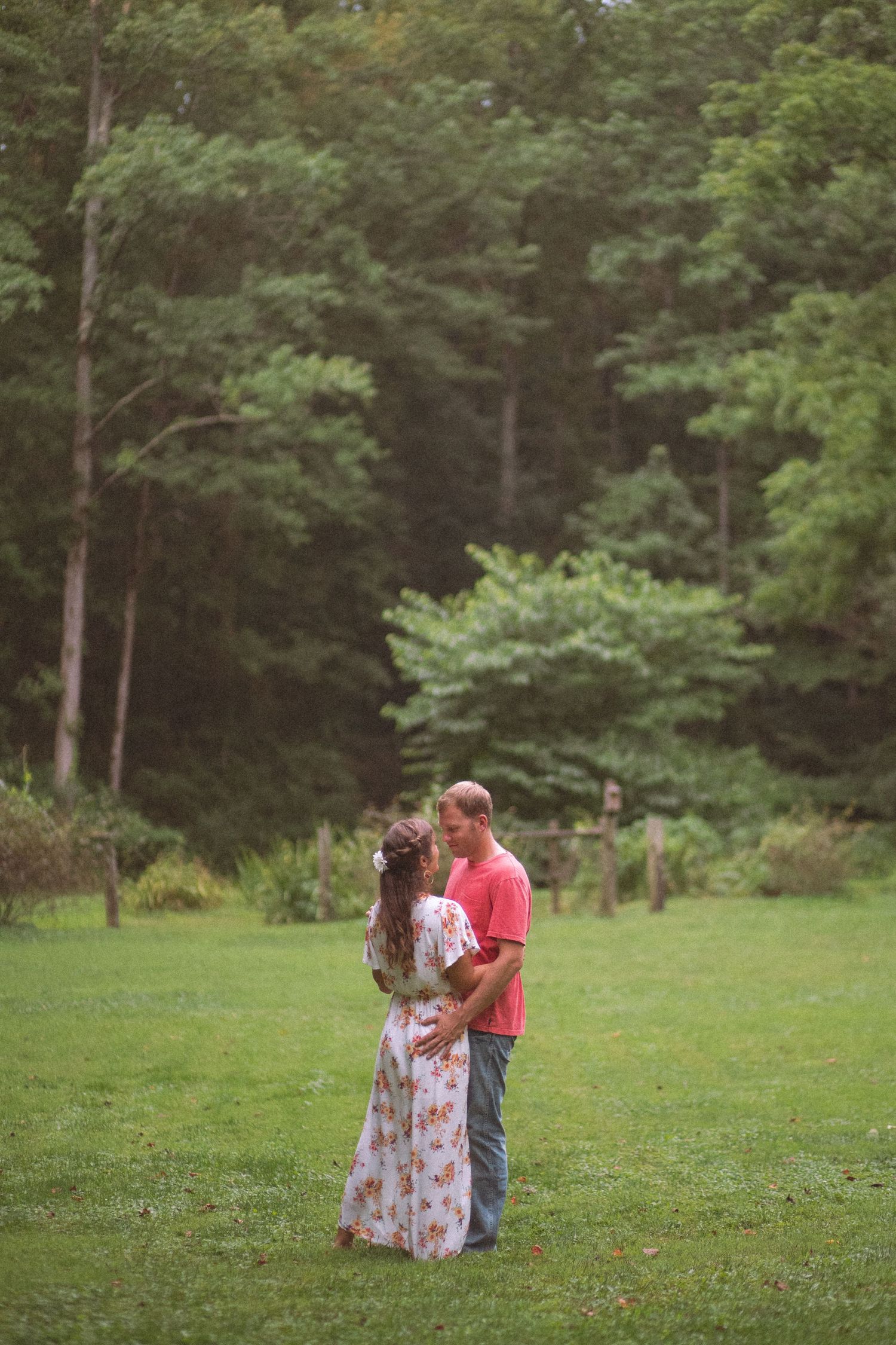 Couple embracing tenderly in a green meadow surrounded by tall trees in a forest clearing.