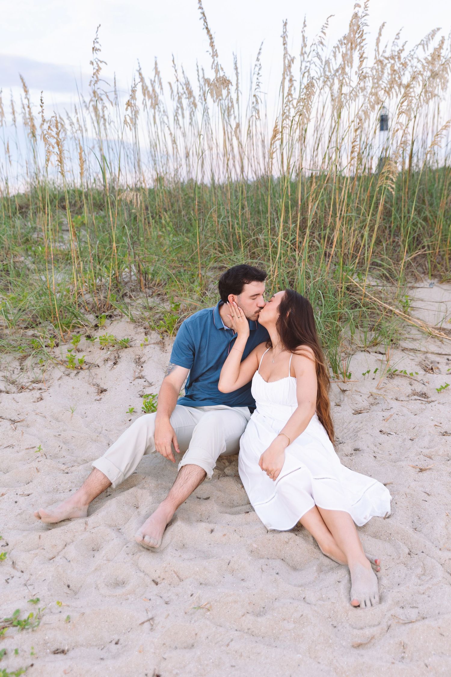 Romantic couple kissing while sitting on sandy beach dunes surrounded by tall sea oats grass.