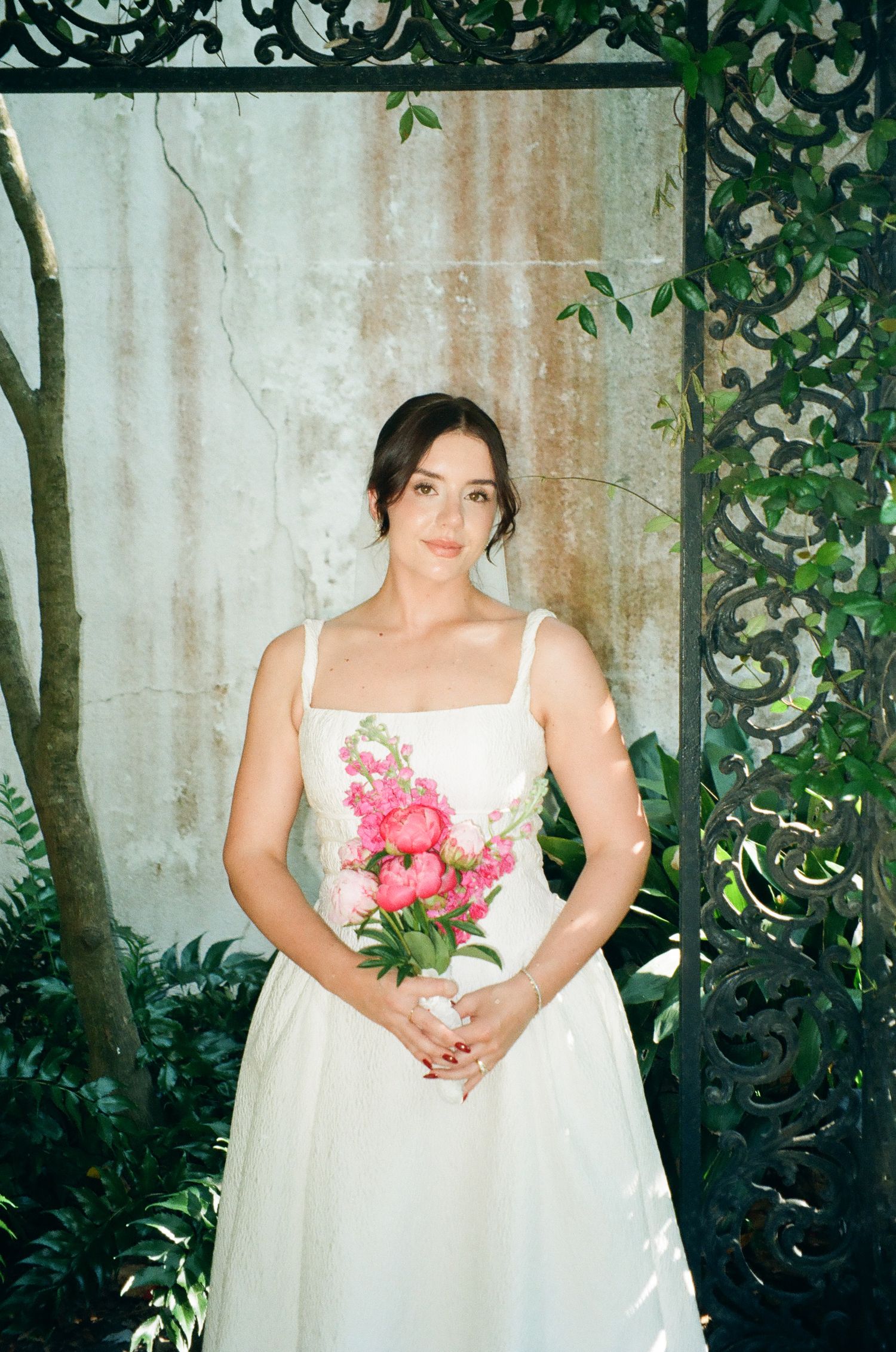 Bride in a white gown with floral embroidery holds a pink peony bouquet framed by ornate iron gates.