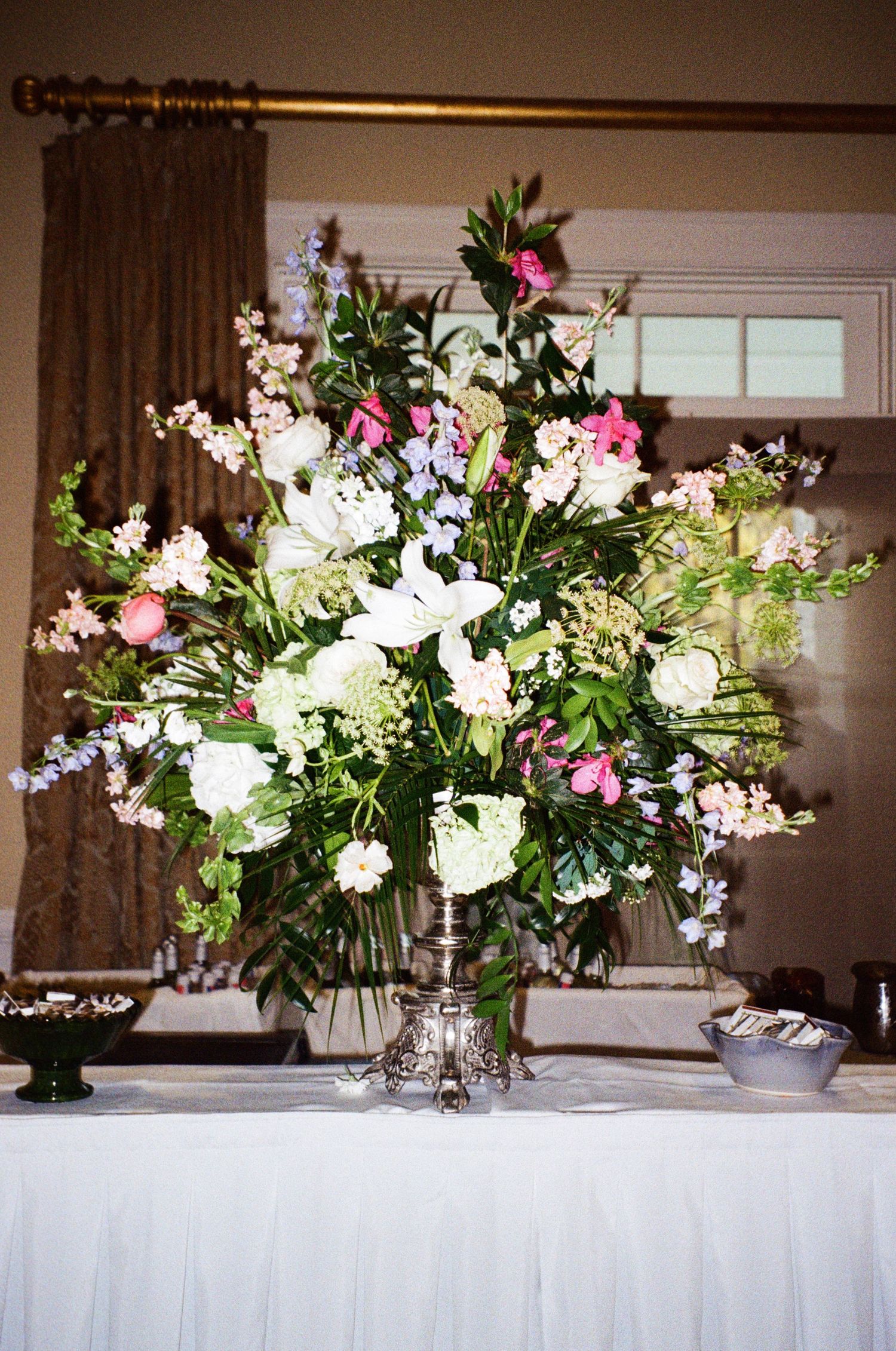 Elegant large floral centerpiece with white lilies, pink blooms, and greenery in a silver vase on a draped table.