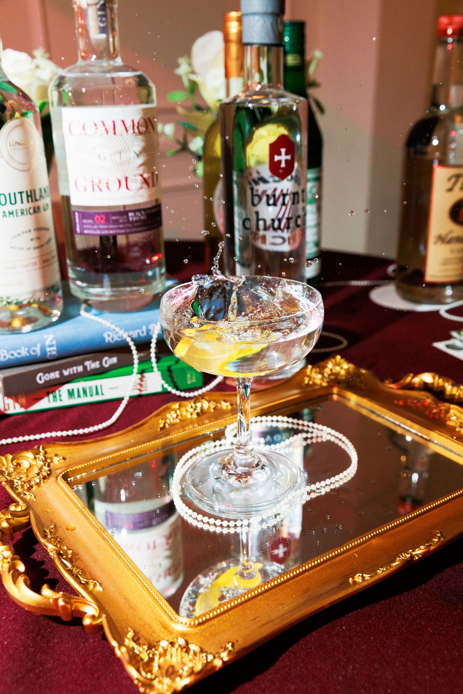 Elegant cocktail splashing in coupe glass on gold mirrored tray with gin bottles including Common Grounds and Bux Chance.