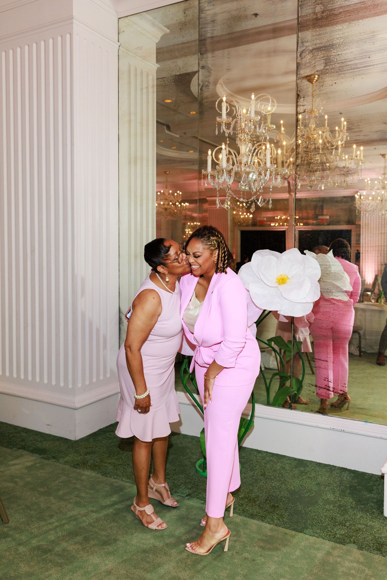 Two women in pink outfits share a kiss near a mirror at an elegant event venue with chandelier.