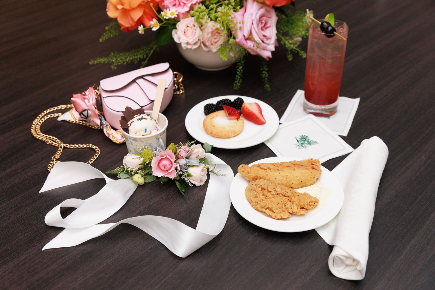 Elegant table setting with floral arrangement, pink handbag, cookies, fruit, and a red cocktail on dark wood.