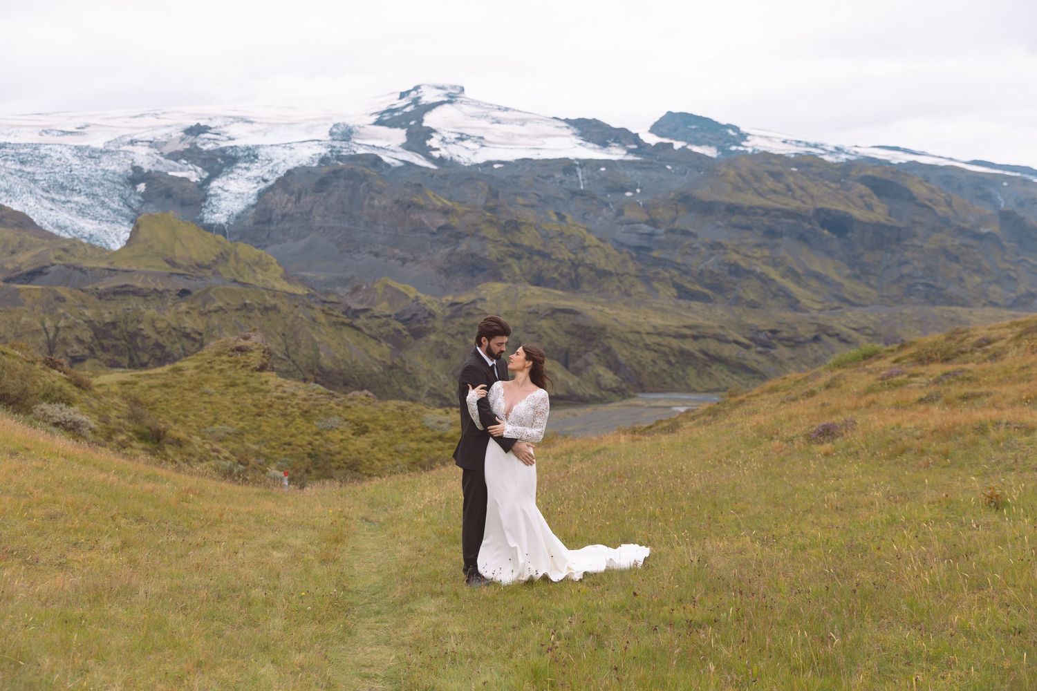 Bride and groom embracing on green Icelandic hillside with breathtaking snow-capped glacier mountains behind them.