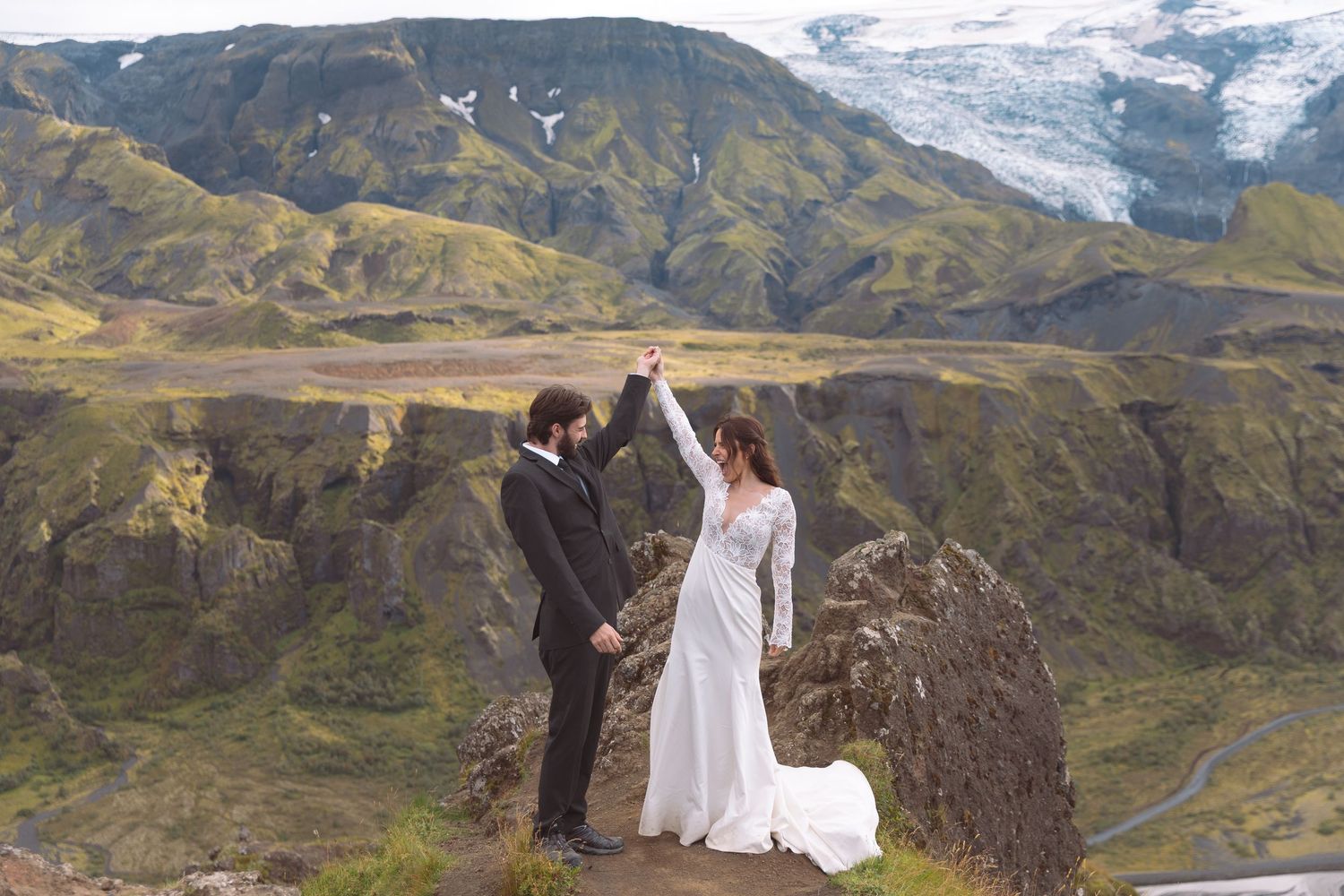 Couple celebrates their Iceland elopement raising hands triumphantly with a glacier and green mountains behind.