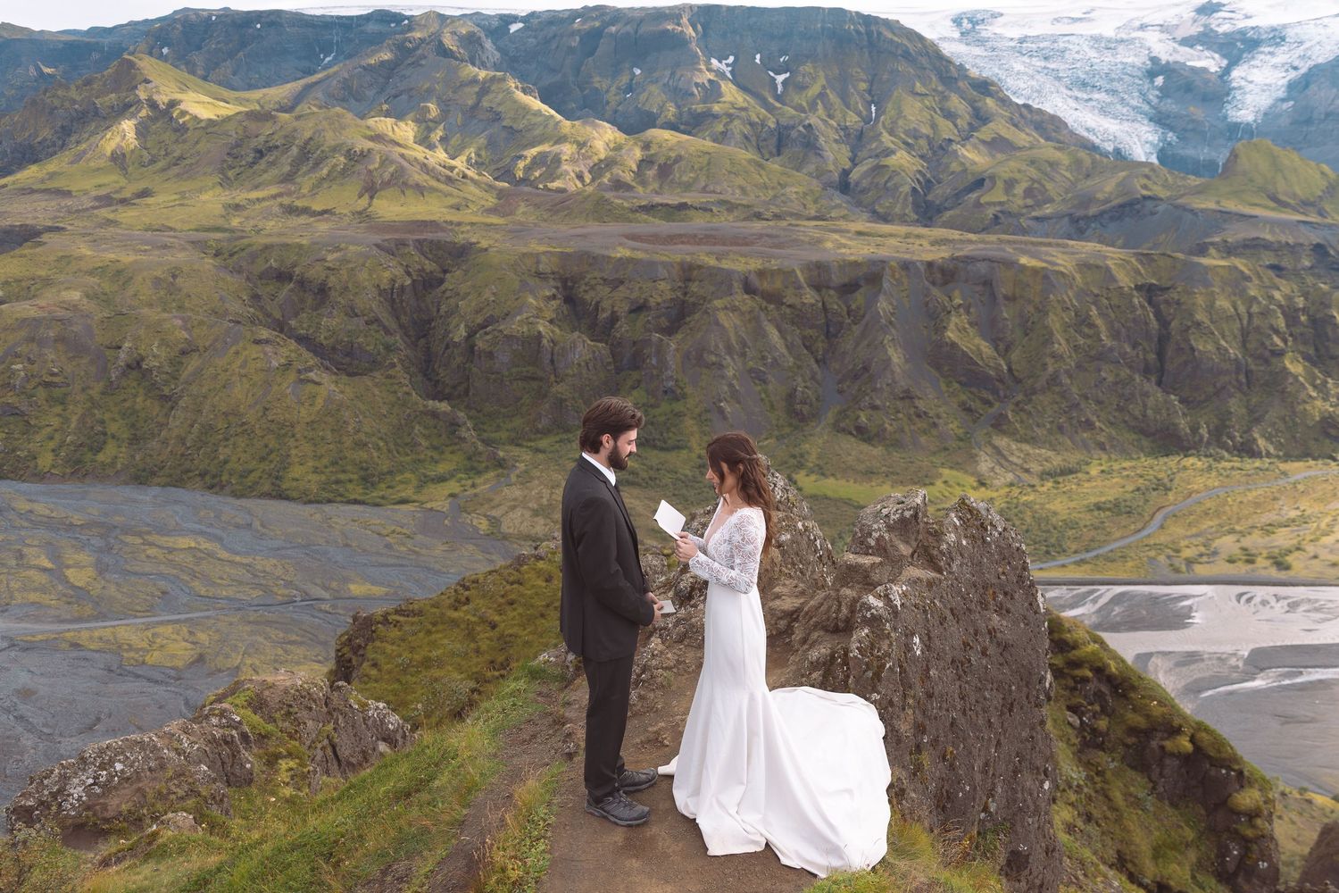 Couple exchanges wedding vows on dramatic Icelandic mountain summit with glacier and green valleys below.