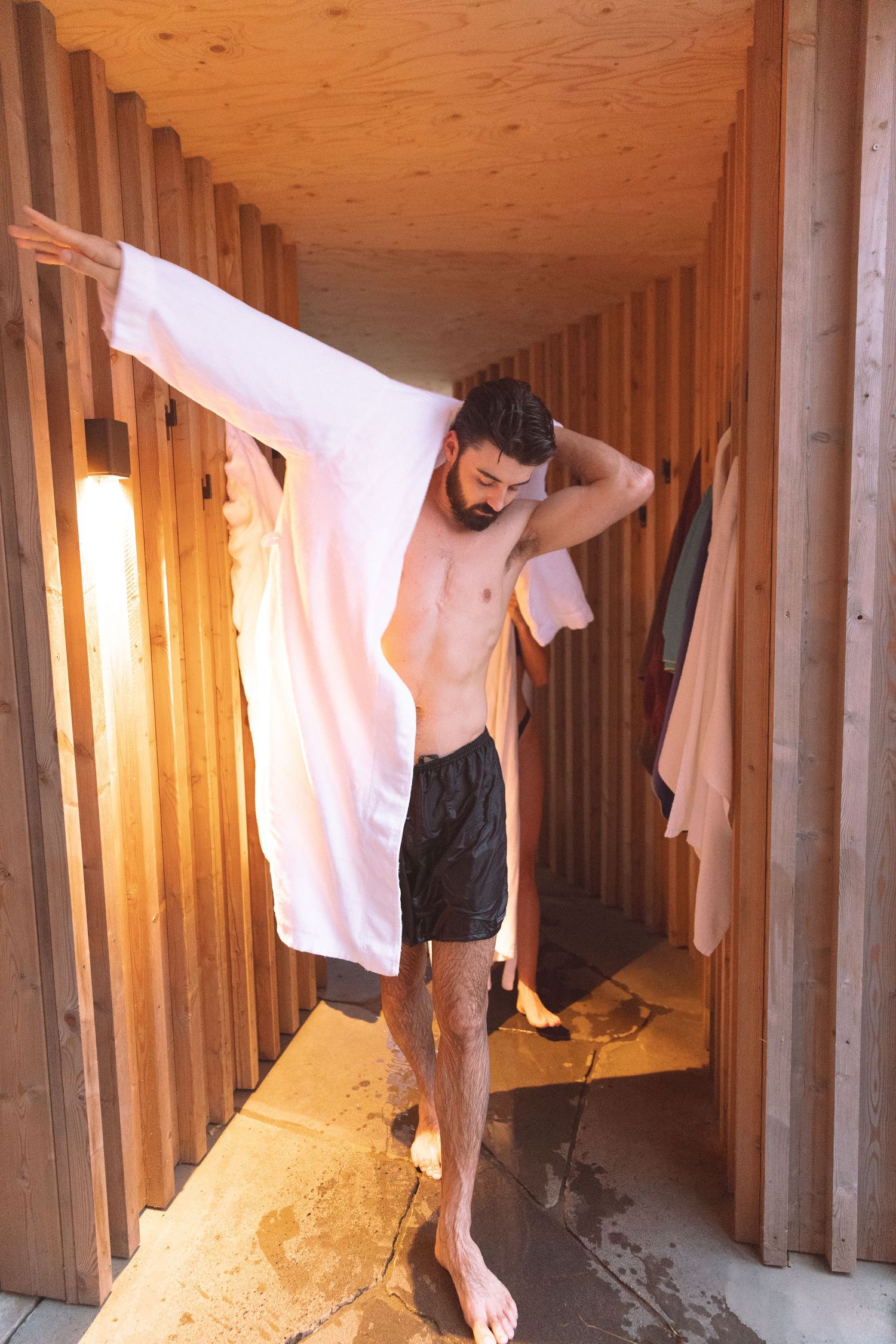 Bearded man in black shorts drying off with white towel in wooden sauna changing room.