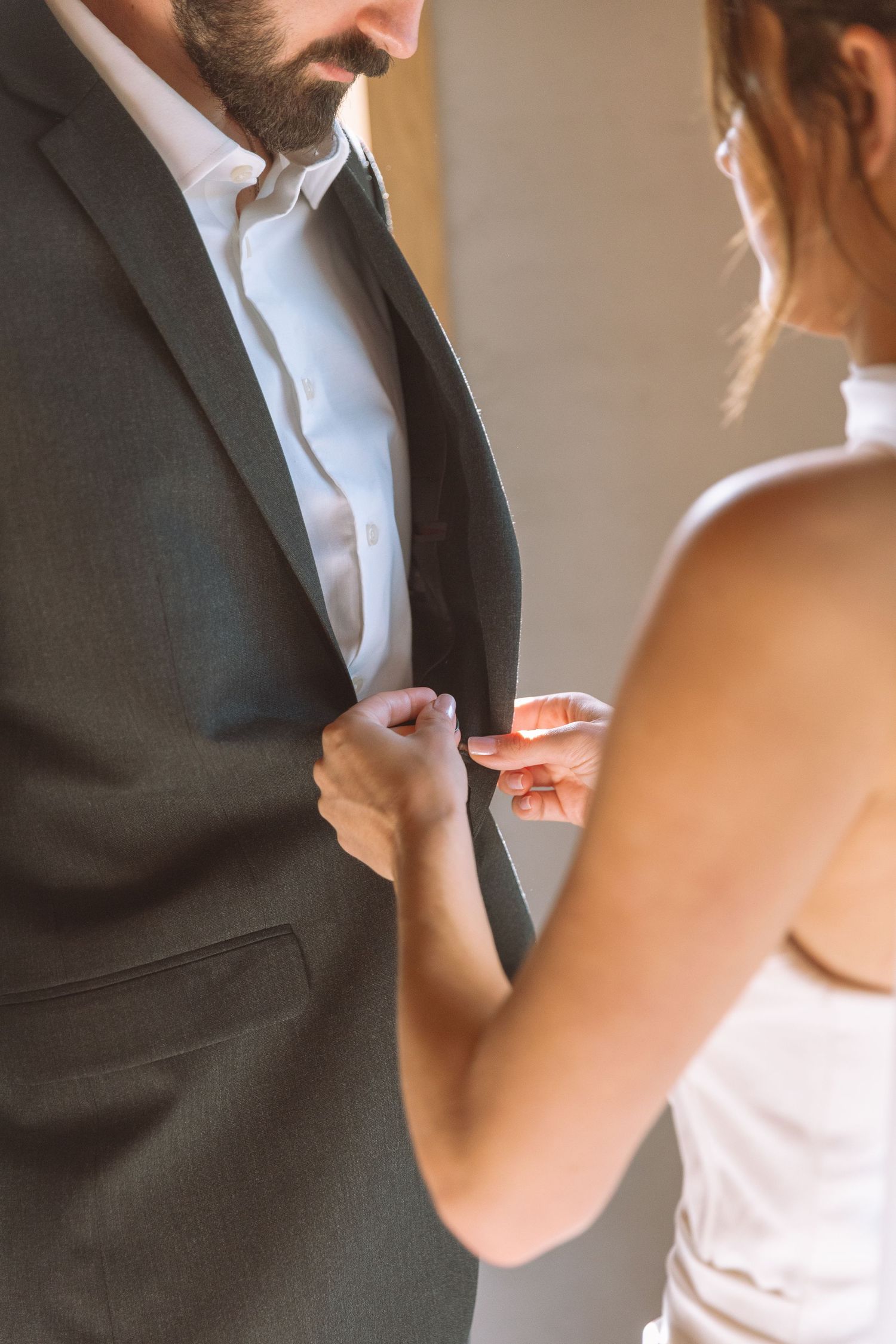Bride adjusting grooms dark suit jacket lapel in a softly lit intimate setting.