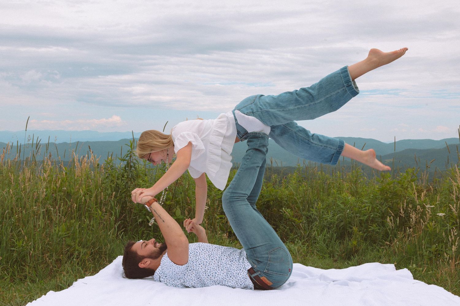 Couple doing acro yoga on a hilltop meadow, man lying on back lifting woman in the air.