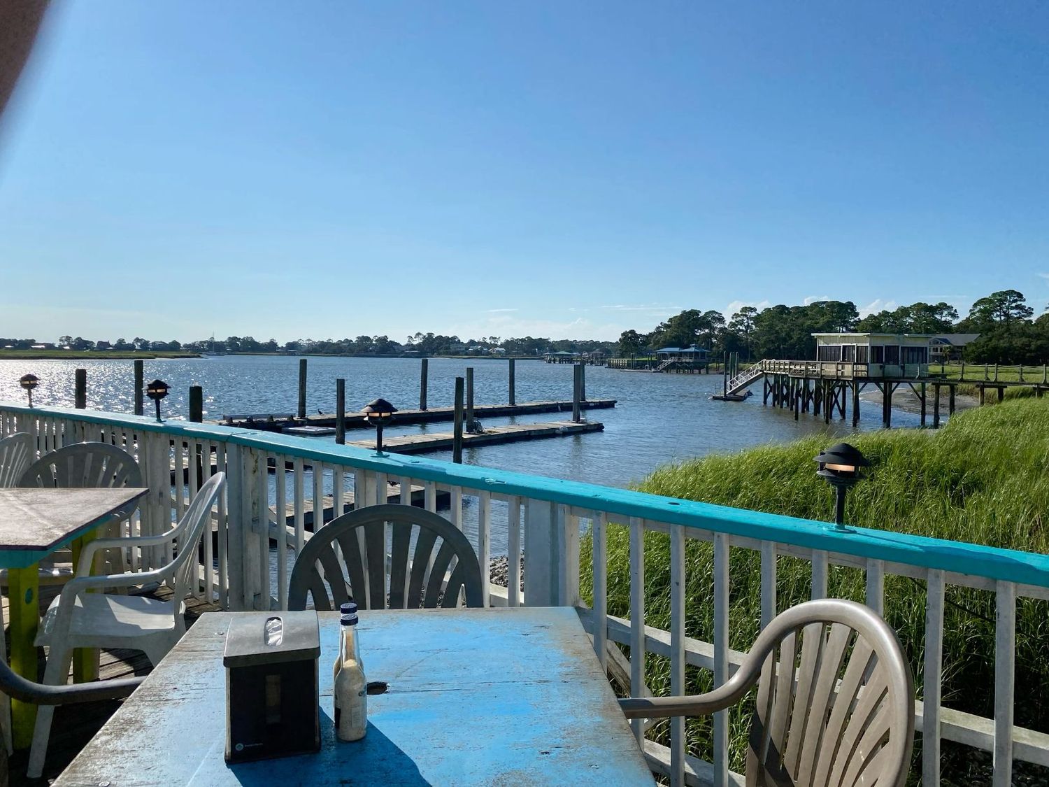 Waterfront restaurant patio with blue table and white chairs overlooking a calm bay with boat docks.